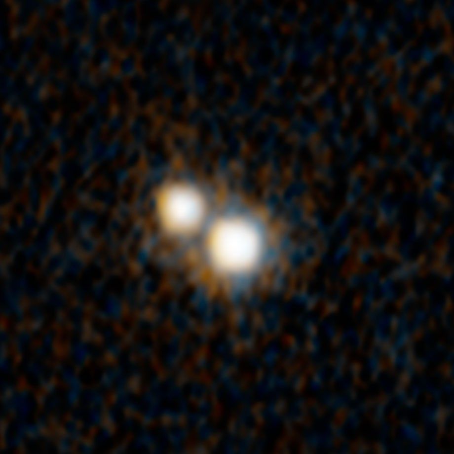 A Hubble Space Telescope photograph of two, closely spaced, white objects, one above and to the left of the other. Each is surrounded by a diffuse orange ring with small orange rays and set against a black background with very faint brownish orange splotches.