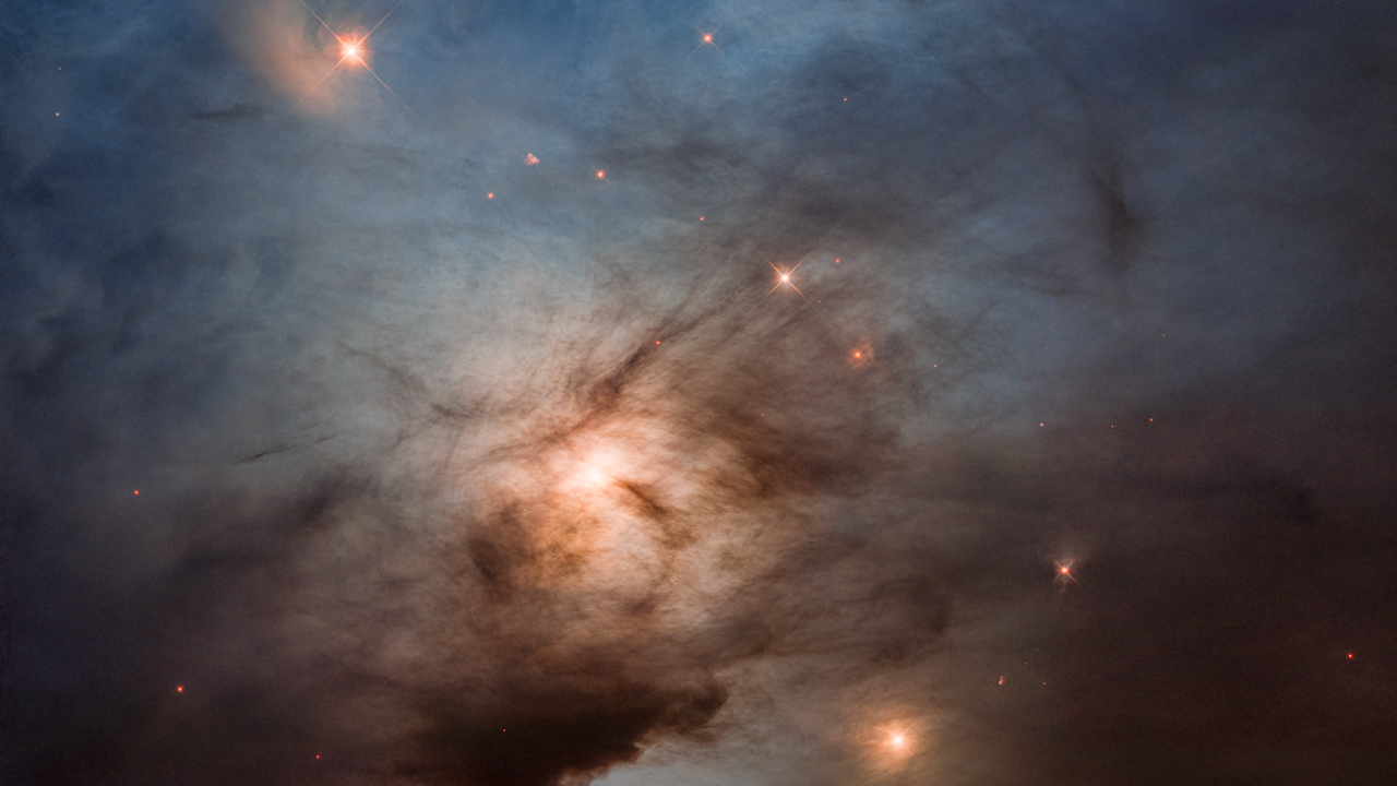 A vertical image with colors ranging from blue at the top to golden in the middle and red at the bottom. At the top, a bright blue star is illuminating surrounding clouds of gas, making the top third of the image appear blue. Below the bright star, a couple dozen fainter stars shine yellow. One of them has a fuzzy, golden arc to its left. At the center of the image, a brighter yellow star illuminates surrounding gas. It is partially obscured by dark streaks and clouds of dust, making it resemble the moon on a cloudy evening. The bottom of the image is noticeably darker than the rest, with the exception of a dramatic splash of red almost like a bug splat on a windshield. A handful of faint, red stars also appear there.