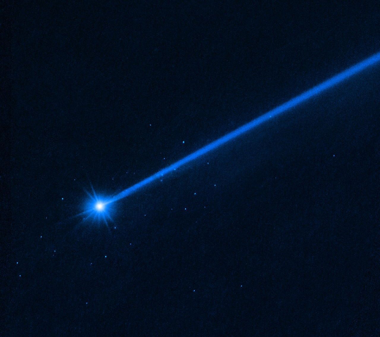 Against a black background, the bright white object at lower left is the asteroid Dimorphos. It has a bluish dust tail extending diagonally to the upper right. A cluster of blue dots surround the asteroid. These are boulders that were knocked off the asteroid when, on September 26, 2022, NASA deliberately slammed the half-ton DART impactor spacecraft into the asteroid as a test of what it would take to deflect some future asteroid from hitting Earth. Hubble photographed the slow-moving boulders with the Wide Field Camera 3 in December 2022. The color results from assigning a blue hue to the monochromatic (grayscale) image.