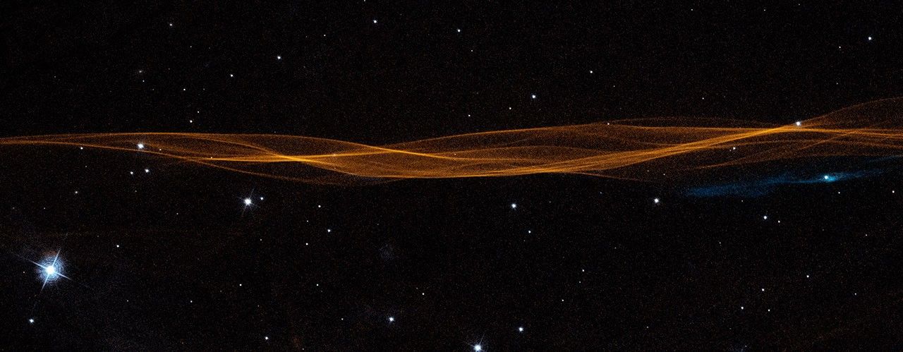 A horizontal, undulating linear structure resembling overlapping strings of yarn extends edge-to-edge across the frame, but takes up only  a quarter of the width at its widest point. Mostly orange with some blue, these are filaments of glowing gasses, which resemble lines in a wrinkled bedsheet.