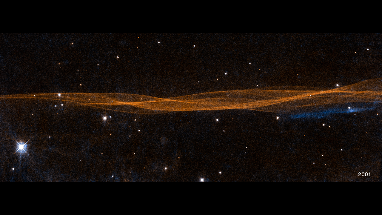 A horizontal, undulating linear structure resembling overlapping strings of yarn extends edge-to-edge across the frame, but takes up only a quarter of the width at its widest point. Mostly orange with some blue, these are filaments of glowing gasses, which resemble lines in a wrinkled bedsheet. The WFPC2 image observation date "2001" is in the bottom right corner.