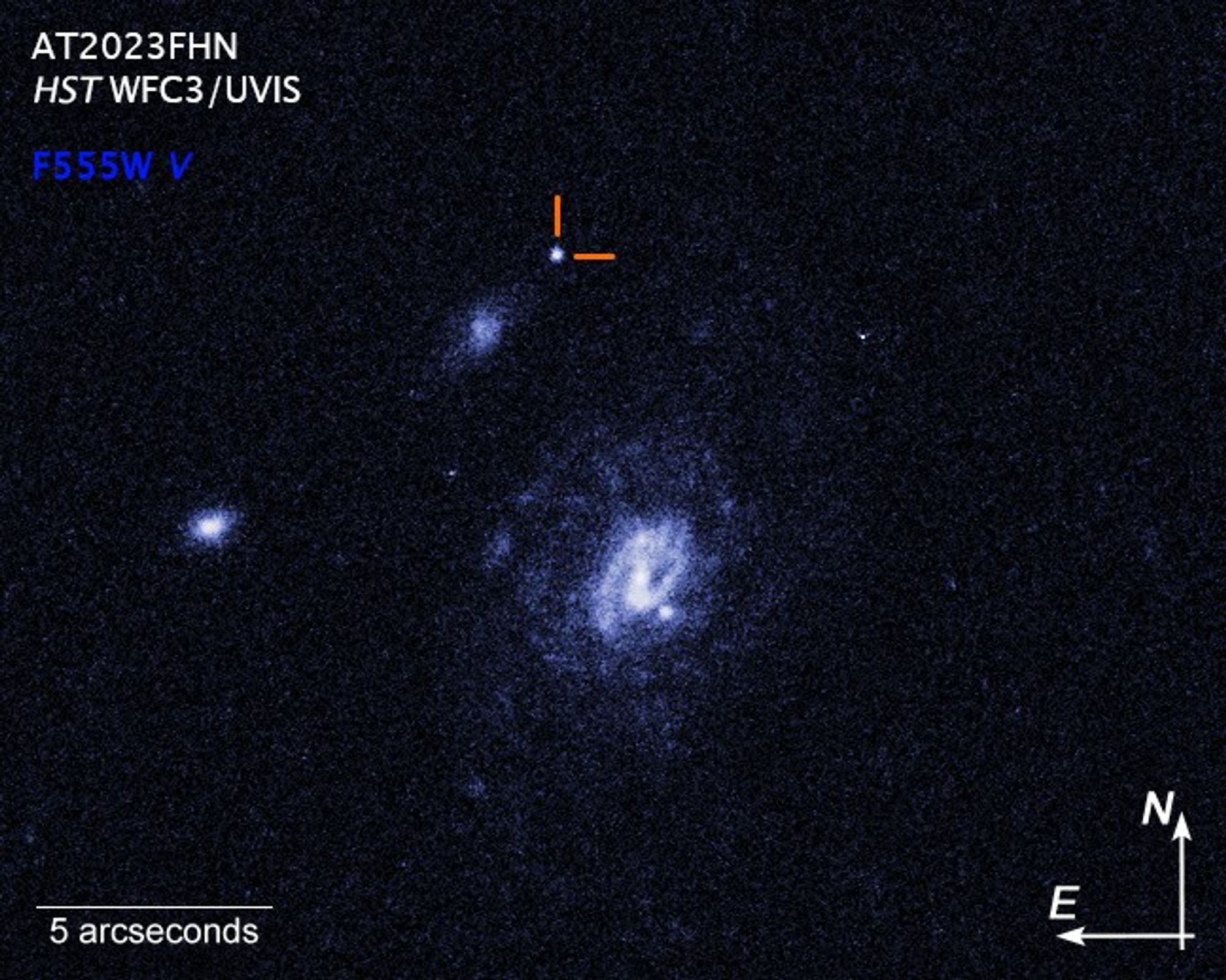 Image titled “AT2023fhn HST WFC3/UVIS” with color key, scale bar and compass arrows shows three galaxies against the velvet-black backdrop of space. The largest is the white and blue spiral-shaped galaxy at image center. Two smaller galaxies are whitish patches toward the left. A curious white spot near the top of the image is the brilliant glow from some unknown object that exploded, but is not associated with any of the galaxies. A color key showing the Hubble instrument filter used to create the image is provided at top left below, the image title. The assigned filter color is blue: F555W V. At bottom right is a white scale bar labeled “5 arcseconds.” At lower right are white compass arrows indicating the orientation of the image on the sky. The east arrow points in the 9 o’clock direction. The north arrow points towards 12 o’clock.