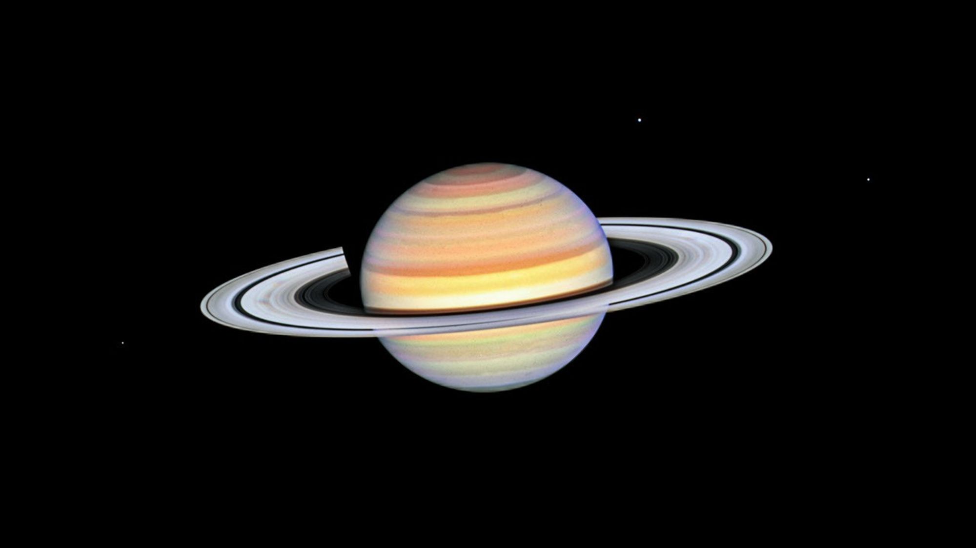 Saturn with bright white rings and multi-colored main sphere. Spoke features on the left and right sides of the rings appear like faint gray smudges against the rings' bright backdrop, about midway from the planet to the rings' outer edge. Above the rings plane, the planet's bands are shades of red, orange and yellow, with bright white nearer the equator. The background of space is black.