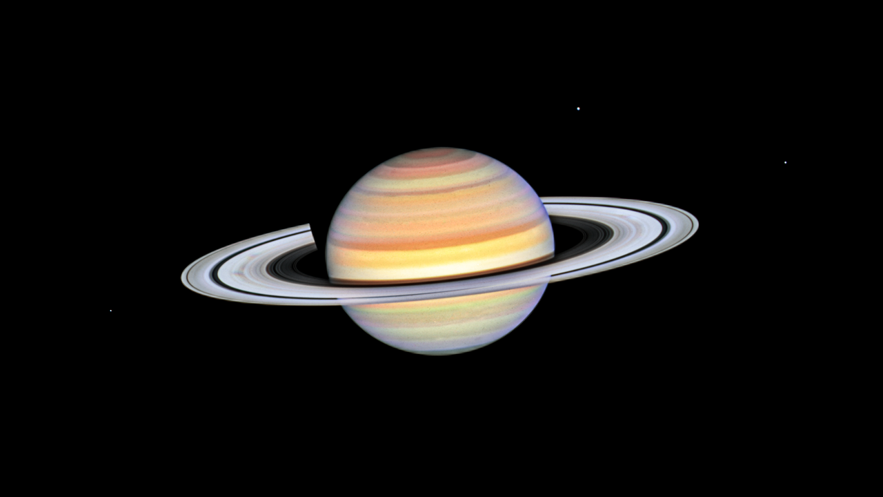 Saturn with bright white rings and multi-colored main sphere. Spoke features on the left and right sides of the rings appear like faint gray smudges against the rings' bright backdrop, about midway from the planet to the rings' outer edge. Above the rings plane, the planet's bands are shades of red, orange and yellow, with bright white nearer the equator. The background of space is black.