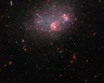 A collection of stars and galaxies fill the scene against a dark background. The image is dominated by a dense collection of stars which make up the irregular galaxy UGC 8091. The stars span a variety of colors, including blue and orange, with patches of blue occupying the central part of the galaxy. There are also visible circular regions of red/pink gas within the galaxy.