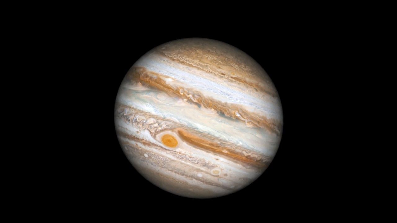 A Hubble Space Telescope image of the giant planet Jupiter, on the black background of space. The planet is banded in stripes of brownish orange, light gray, soft yellow, and shades of cream. It's punctuated with many large storms and white clouds. The largest storm, the Great Red Spot, appears near the left bottom third of the planet.