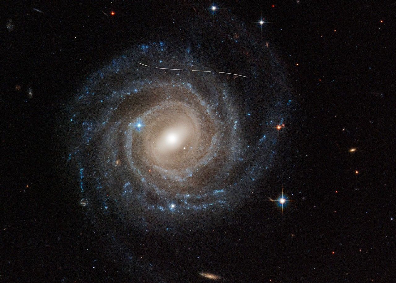 This is a Hubble Space Telescope image of the barred spiral galaxy UGC 12158. The majestic galaxy has a pinwheel shape made up of bright blue stars wound around a yellow-white hub of central stars. The hub has a slash of stars across it, called a bar. The galaxy is tilted face-on to our view from Earth. A slightly s-shaped white line across the top is a Hubble image is of an asteroid streaking across Hubble's view. It looks dashed because the image is a combination of several exposures of the asteroid flying by like a race car.