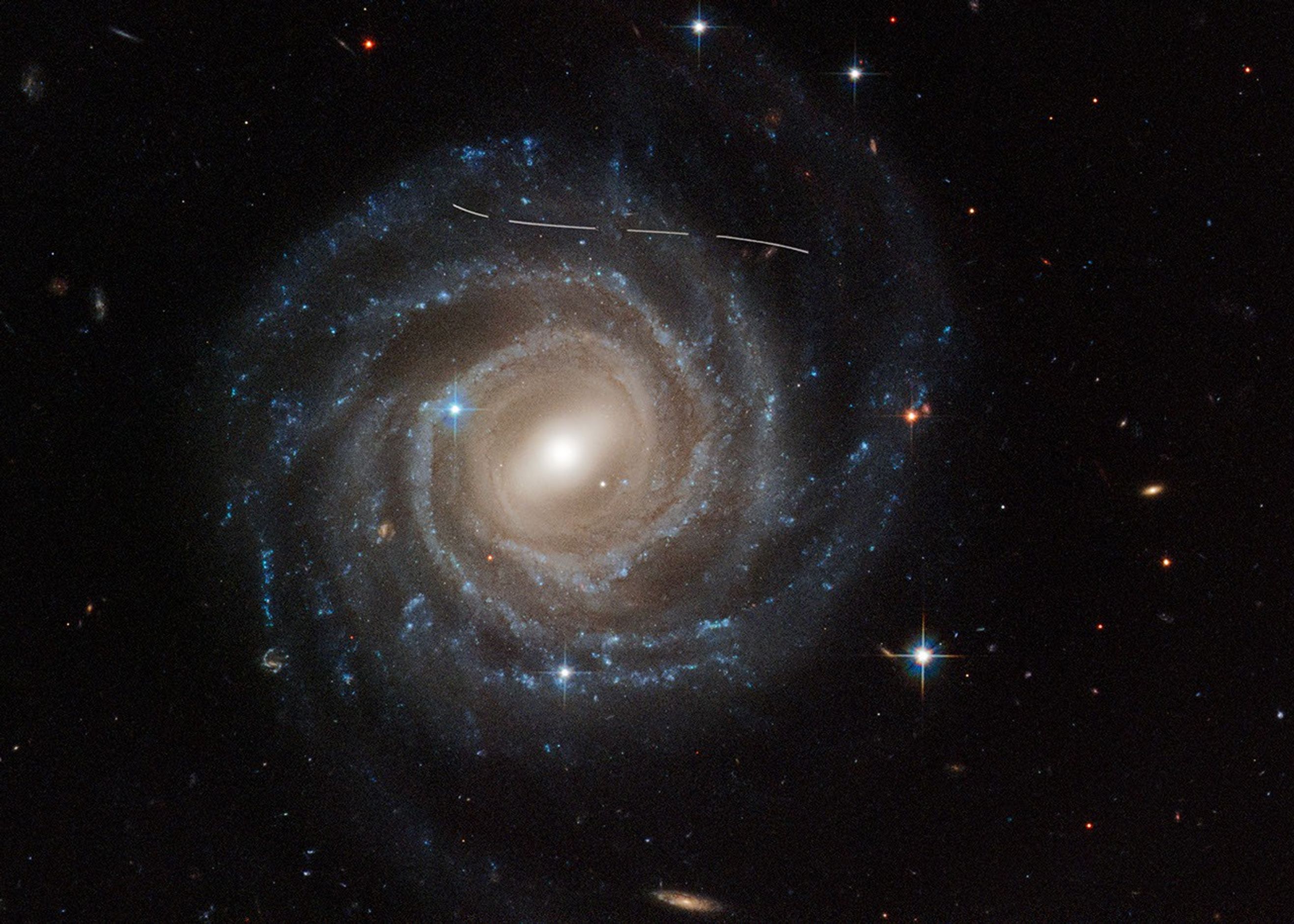 This is a Hubble Space Telescope image of the barred spiral galaxy UGC 12158. The majestic galaxy has a pinwheel shape made up of bright blue stars wound around a yellow-white hub of central stars. The hub has a slash of stars across it, called a bar. The galaxy is tilted face-on to our view from Earth. A slightly s-shaped white line across the top is a Hubble image is of an asteroid streaking across Hubble's view. It looks dashed because the image is a combination of several exposures of the asteroid flying by like a race car.