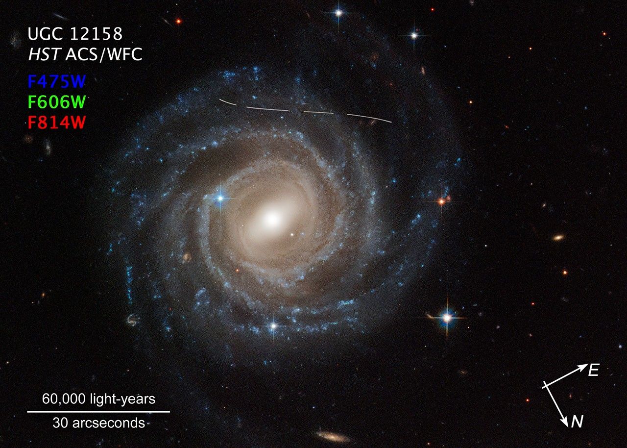 Annotated image of barred spiral galaxy UGC 12158 against the black background of space, with compass arrows, a scale bar, and color key for reference. The galaxy has a pinwheel shape made up of bright blue stars wound around a yellow-white hub of central stars. The hub has a slash of stars across it, called a bar. The galaxy is tilted face-on to our view from Earth. A slightly s-shaped white line across the top is a Hubble image is of an asteroid streaking across Hubble's view. It looks dashed because the image is a combination of several exposures of the asteroid flying by like a race car. White text at top left reads "UGC 12158." White text below reads "HST ACS/WFC." Near top left, color key consisting of three lines reads: "F475" in blue, "F606" in green, and "F814W" in red. At bottom left corner is a scale bar labeled "60,000 light-years" over "30 arcseconds." At bottom right corner, the "E" compass arrow points towards the 2 o'clock position. The "N" compass arrow points towards the 5 o'clock position.