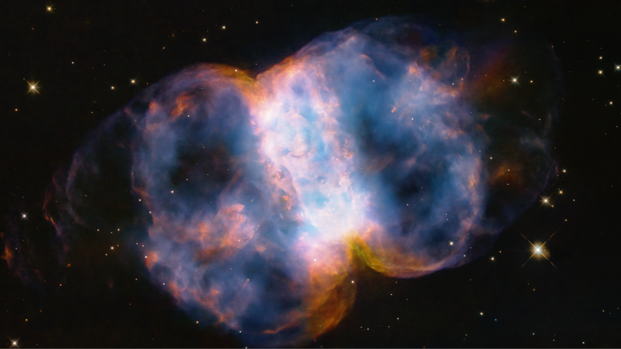 M76_1280x720 - Taking up most of the image, is a multi-colored nebula appearing as two translucent orbs attached by a white band.