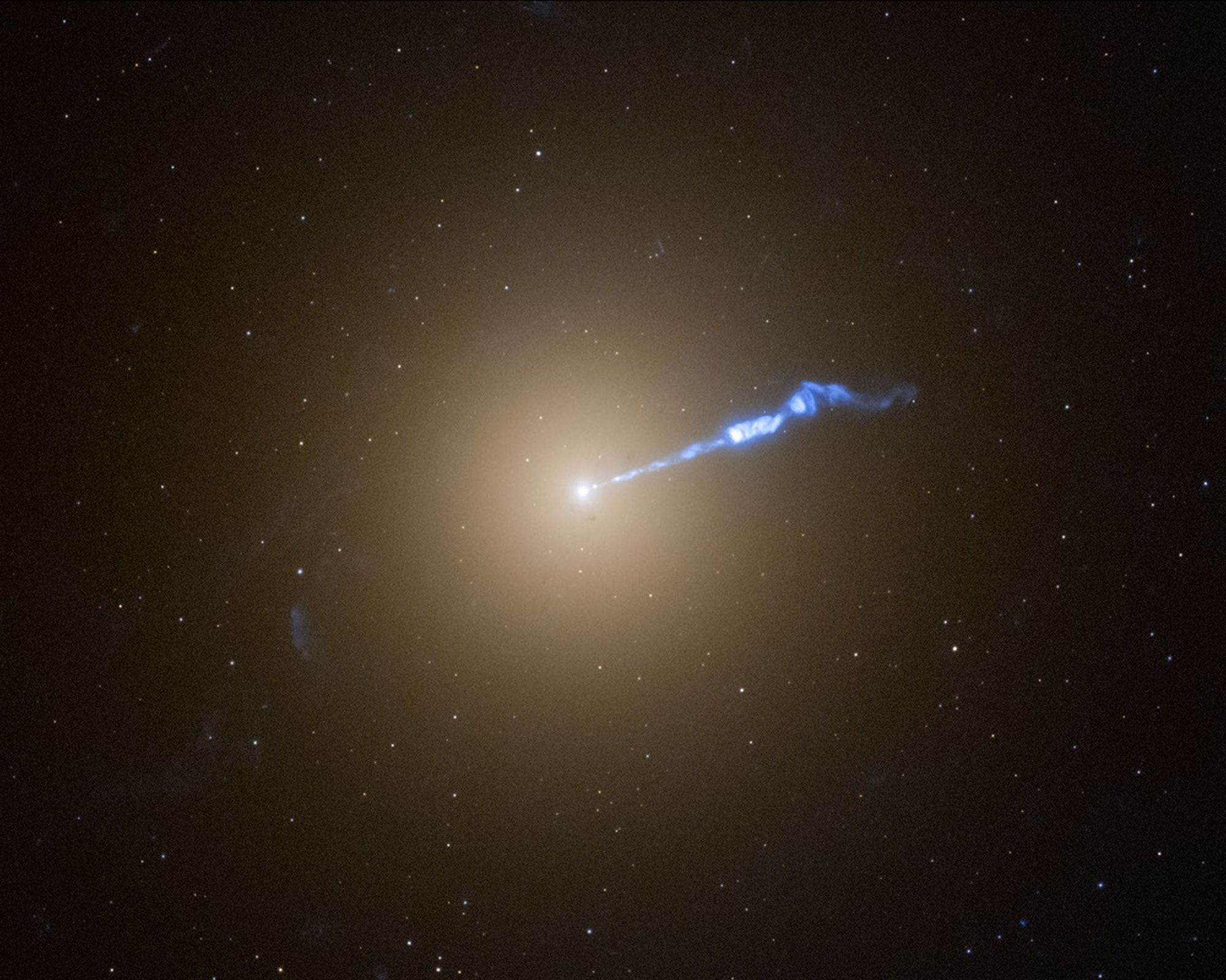 A Hubble  photo of galaxy M87, which resembles a translucent, fuzzy white cotton ball. The brightness decreases gradually out in all directions from a bright white point of light at the center. A wavy blue-white jet of material extends from the point-like core outward to the upper right, about halfway across the galaxy. Stars speckle the background.