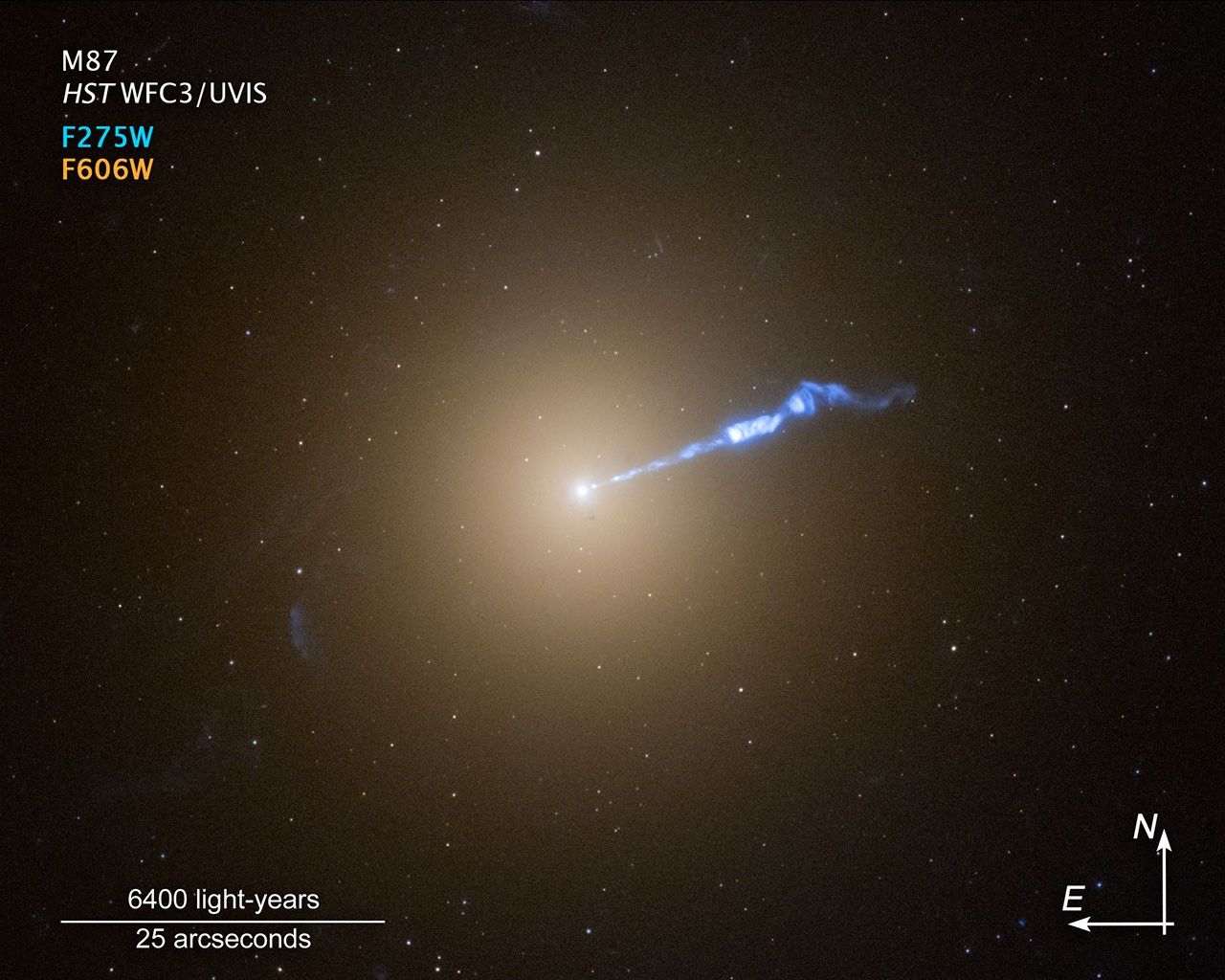 Image titled "M87, HST/WFC3/UVIS" with color key, scale bar and compass arrows on a black background of space. The M87 galaxy resembles a translucent, fuzzy white cotton ball. The brightness decreases gradually out in all directions from a bright white point of light at the center. A wavy blue-white jet of material extends from the point-like core outward to the upper right, about halfway across the galaxy. Stars speckle the background. At upper left is a color key, showing which Hubble Wide Field Camera 3 filters were used to create the image and which color is assigned to each filter: F275W, blue; F606W, orange. A scale bar labeled "6400 light-years over 25 arcseconds" is in the bottom left corner of the image. At the bottom right corner of the image are compass arrows indicating the orientation of the image on the sky. The east arrow points toward 9 o'clock. The north arrow points to the 12 o'clock direction.