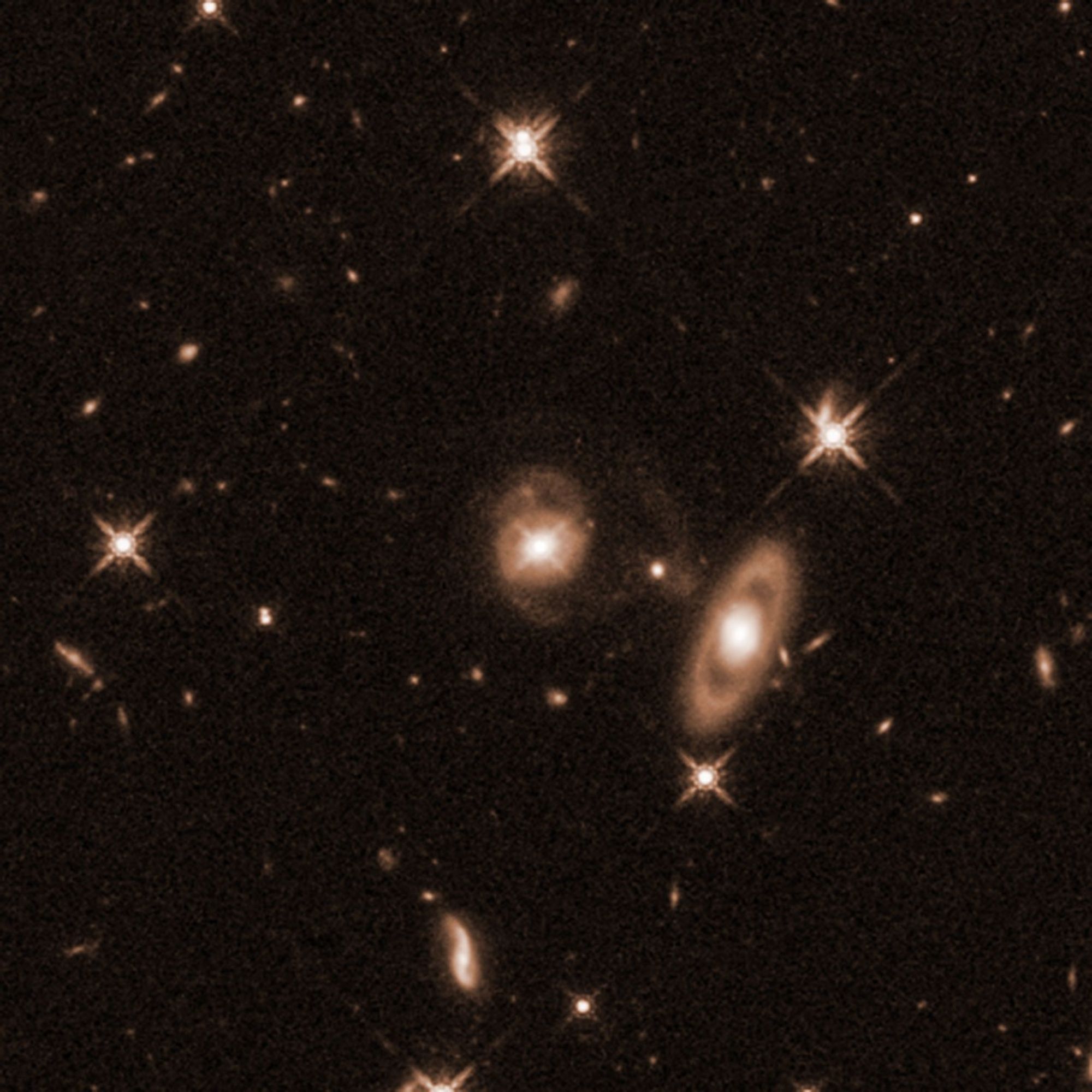 Field of yellow galaxies of various sizes and distances on a black background. Two larger galaxies are prominent. Centered is a galaxy with a bright core and faint spiral arms coming off its top and bottom. To the lower right of the spiral is a ring galaxy with an apparent gap between its bright core and the ring oval of dust and gas surrounding it.