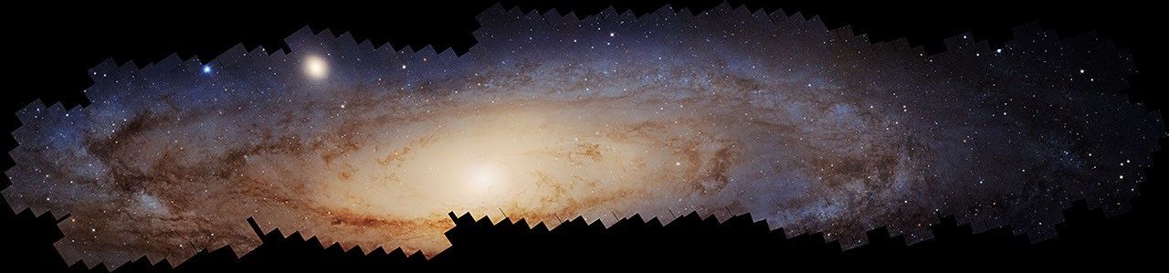 The Andromeda galaxy, a spiral galaxy, spreads across the width. It is tilted nearly edge-on to our line of sight so that it appears as an extreme oval on its side. The borders of the galaxy are jagged because the image is a mosaic of smaller, square images. The outer edges are blue, while the inner two-thirds are yellowish with a bright, central core. Dark, dusty filamentary clouds wrap around the outer half of the galaxy’s disk. At 10 o'clock, a smaller dwarf elliptical galaxy forms a fuzzy, yellow blob. Hubble's sharp vision distinguishes about 200 million stars within the image. The background of space is black. There are what appears to be steps toward the bottom, mainly toward the middle, which indicates where no data were taken.