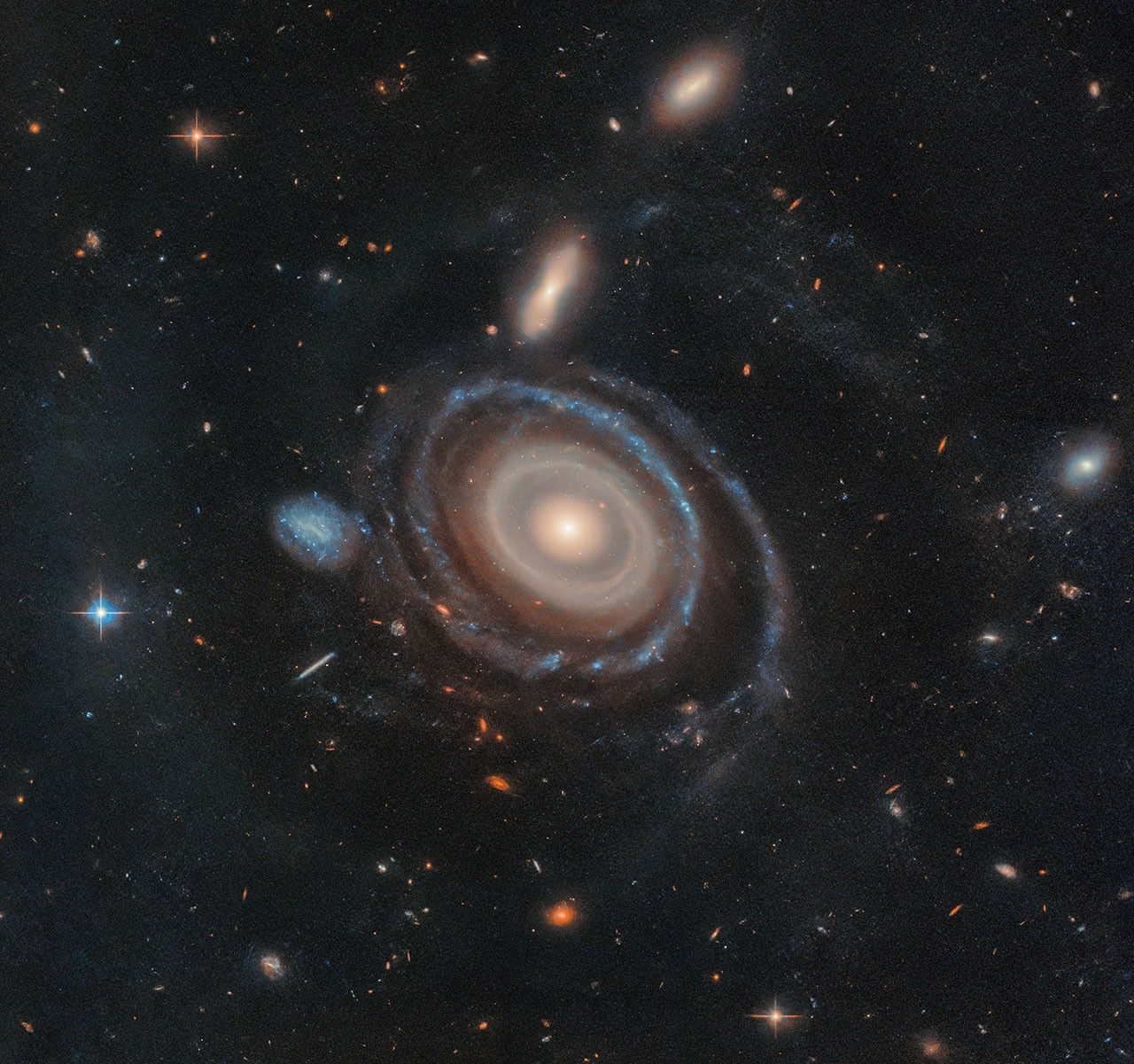 A large galaxy is at center, and a significantly smaller galaxy is to its immediate left. Both are set on the black background of space, which is dotted with a range of galaxies in different shapes and sizes, along with a few foreground stars.