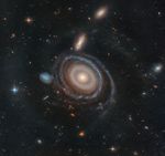 A large galaxy is at center, and a significantly smaller galaxy is to its immediate left. Both are set on the black background of space, which is dotted with a range of galaxies in different shapes and sizes, along with a few foreground stars.