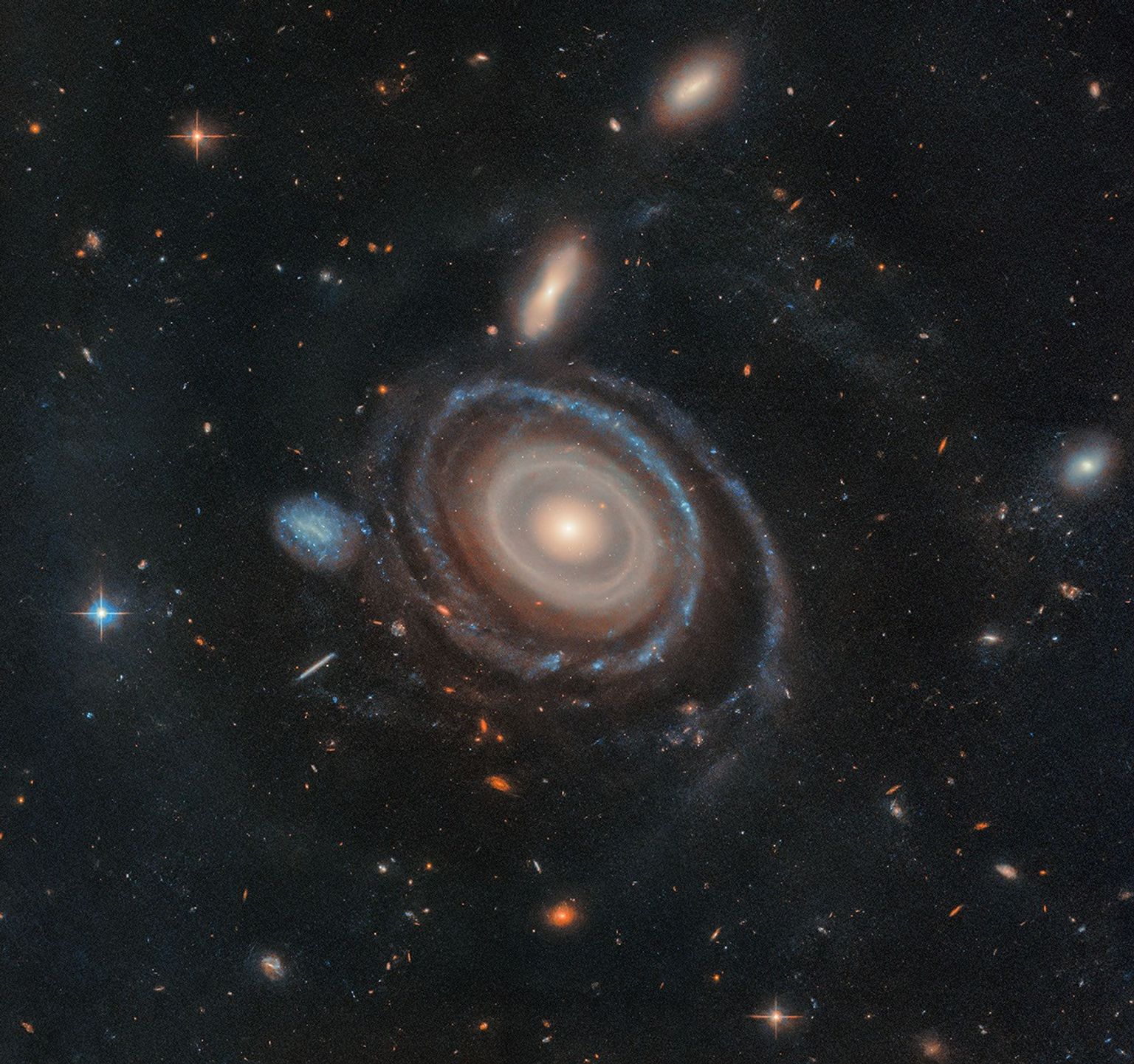 A large galaxy is at center, and a significantly smaller galaxy is to its immediate left. Both are set on the black background of space, which is dotted with a range of galaxies in different shapes and sizes, along with a few foreground stars.