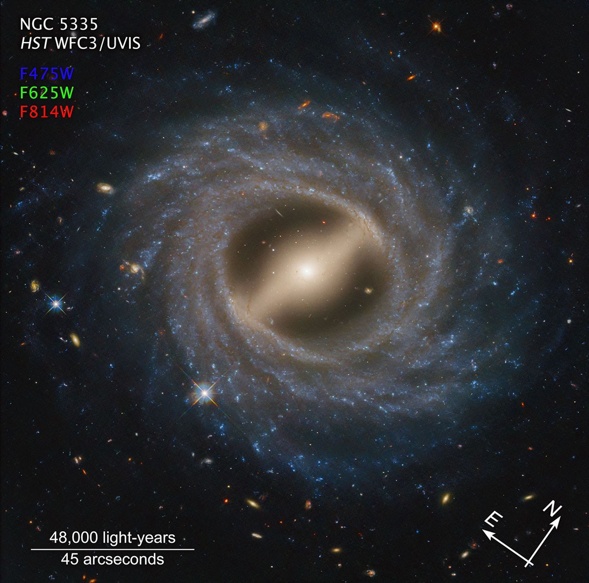Image titled “NGC 5335, HST WFC3/UVIS” at top left with compass arrows and color key at the bottom. Below the name, filters are listed: F475W in blue, F625W in green, and F814W in red. These colors indicate the visible-light color is assigned to each filter. Barred spiral galaxy NGC 5335 takes up the majority of the view. At its center is a milky yellow, flattened oval surrounded by what looks like a bar extending from top left to bottom right. Around this is a thick swath of blue stars speckled with white regions. Multiple arms wrap up and around in a counterclockwise direction. Distant galaxies in an array of colors are speckled throughout the black background. The length of the scale bar at bottom left is about a quarter of the total width. It is labeled 48,000 light-years and 45 arcseconds. At the bottom right are compass arrows indicating the orientation of the image on the sky. The east arrow points toward 10 o’clock. The north arrow points toward 2 o’clock.