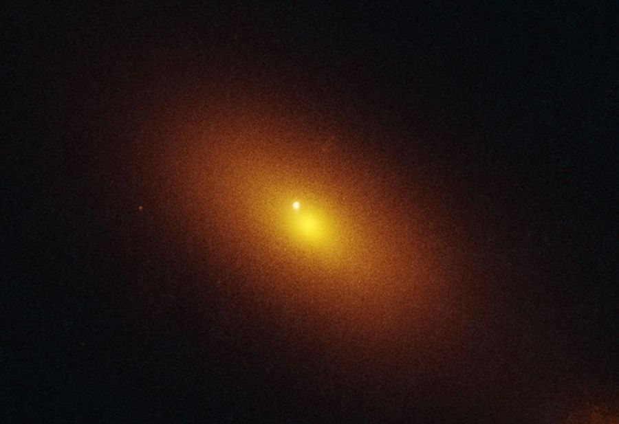 A fuzzy orange-yellow oval occupies the middle fourth of the image. It extends from 10 o’clock to 4 o’clock. A bright white spot is located within the oval but offset to the upper left from the core. An additional smaller, grainy, orange disk is located at lower right. A couple of additional orange points are in the frame. The background of space is black.