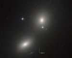 Two elliptical galaxies at upper right and lower left appear as fuzzy white ovals. Two bright stars with prominent diffraction spikes also appear near center of the image. At bottom center is a tiny white dot labeled HLX-1. The background of space is black, and is speckled with tiny white dots.