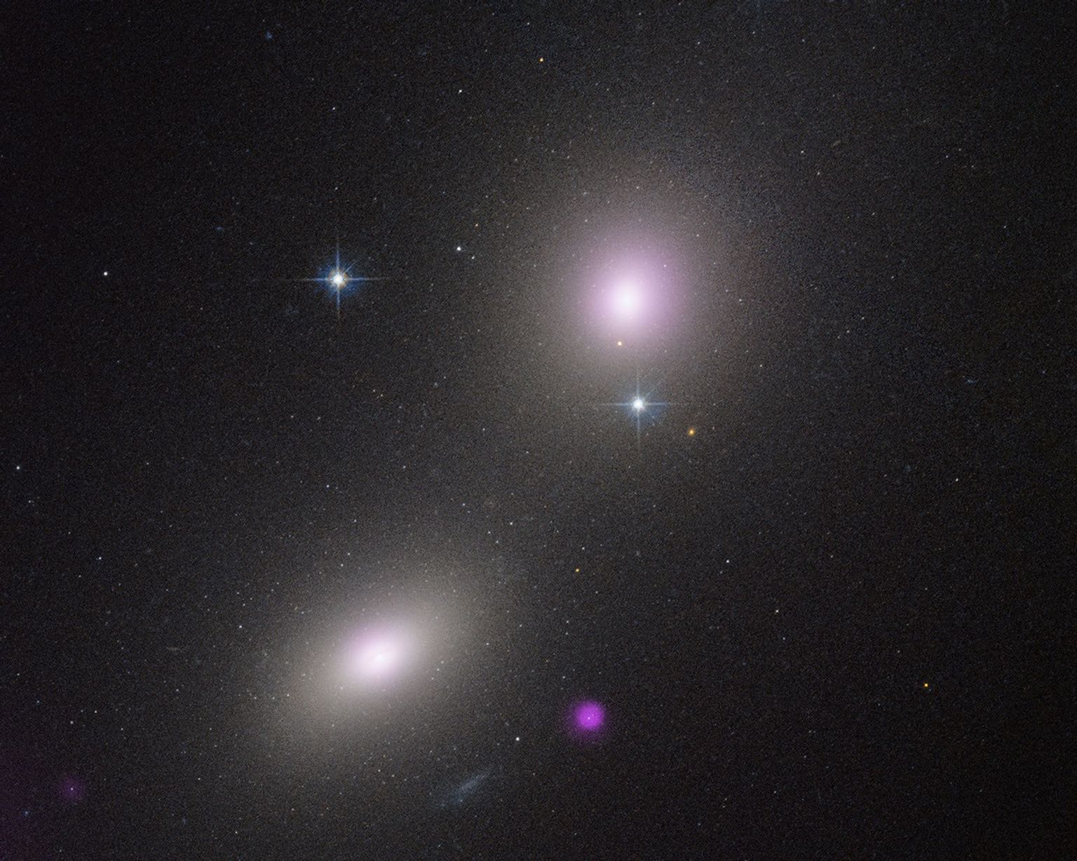 2 Elliptical galaxies appear as fuzzy white oval. At right is a smaller fuzzy purple blob. The background of space is black, speckled with tiny white dots.