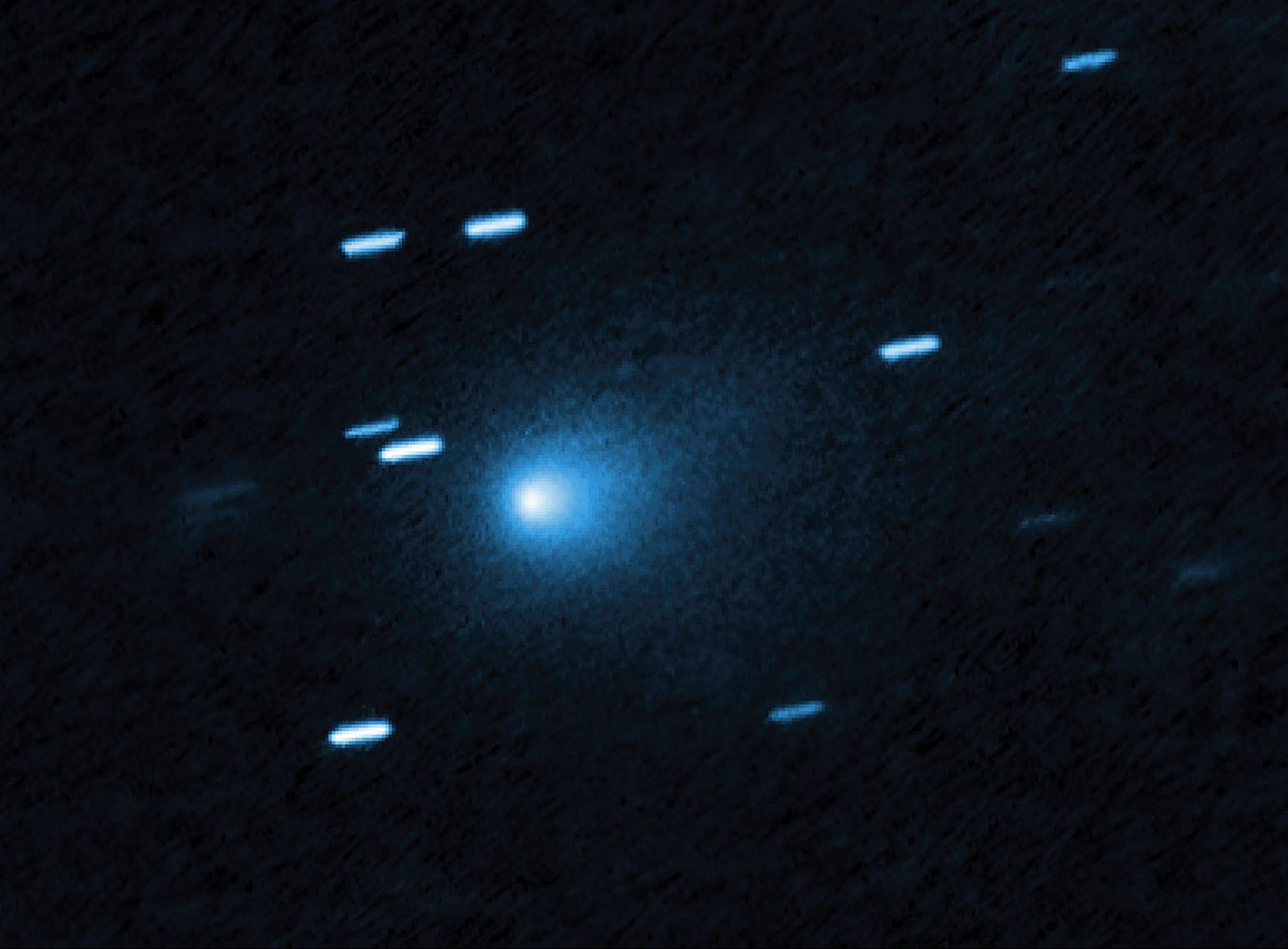 At the center of the image is a comet that appears as a teardrop-shaped bluish cocoon of dust coming off the comet’s solid, icy nucleus and seen against a black background. The comet appears to be heading to the bottom left corner of the image. About a dozen short, light blue diagonal streaks are seen scattered across the image, which are from background stars that appeared to move during the exposure because the telescope was tracking the moving comet.