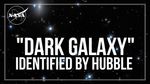 The phrase " 'Dark Galaxy' Identified by Hubble" appears in large white letters.  An outline of the NASA Meatball logo in white appears in the upper left corner. The background is black with a smattering of stars and a few distant galaxies.