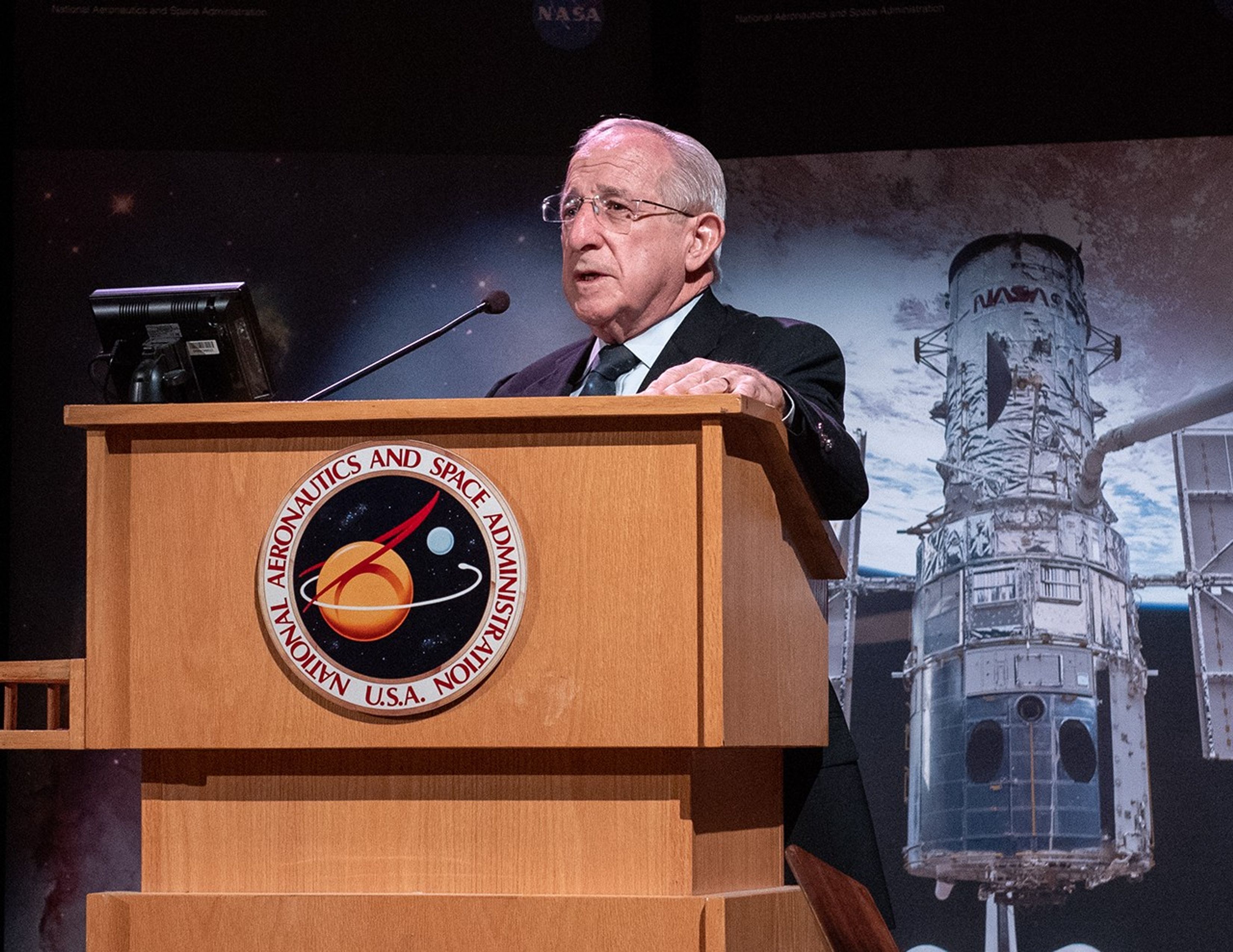 Joseph Rothenberg stands at the podium, addressing the audience, with a banner of the Hubble Space Telescope behind him.