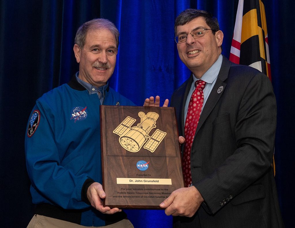 Scolese (right) and Grunsfeld (left) hold a wooden plaque between them with Grunsfeld's name and a gold graphic of Hubble on it.