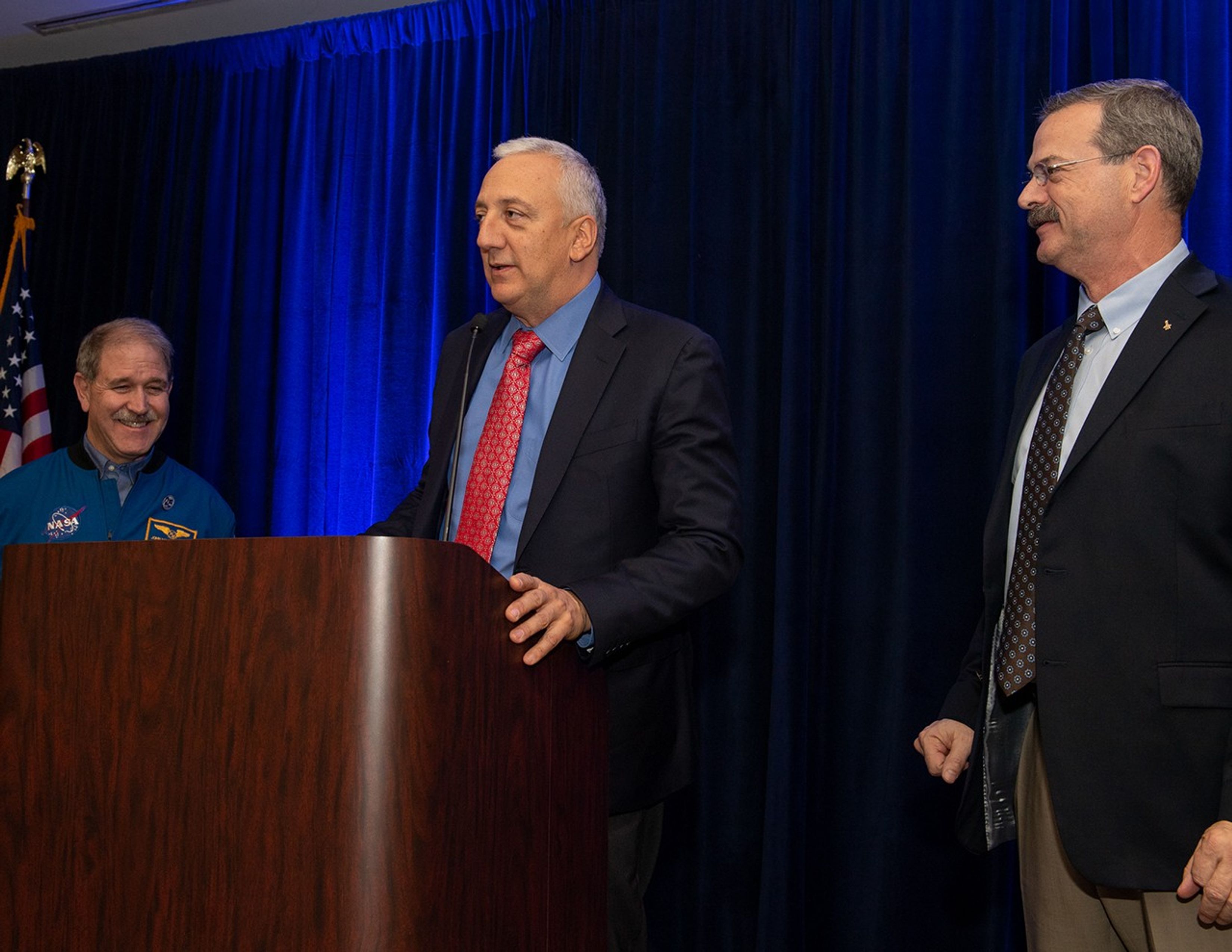 Massimino (center) speaks from the podium while Grunsfeld (left) and Altman (right) watch.