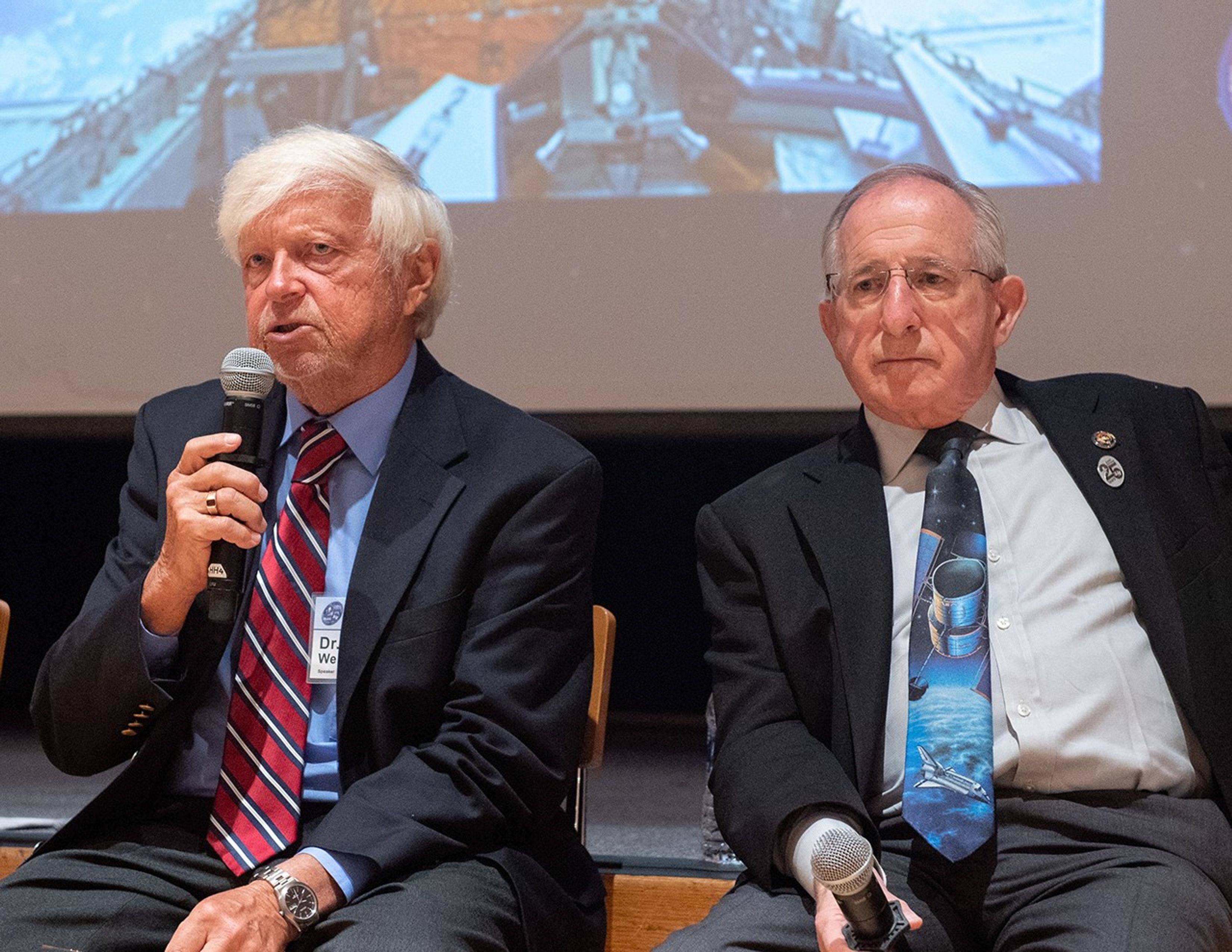 Ed Weiler, left, speaks into a microphone to address the audience, while Joseph Rothenberg, right, also holds a mic.