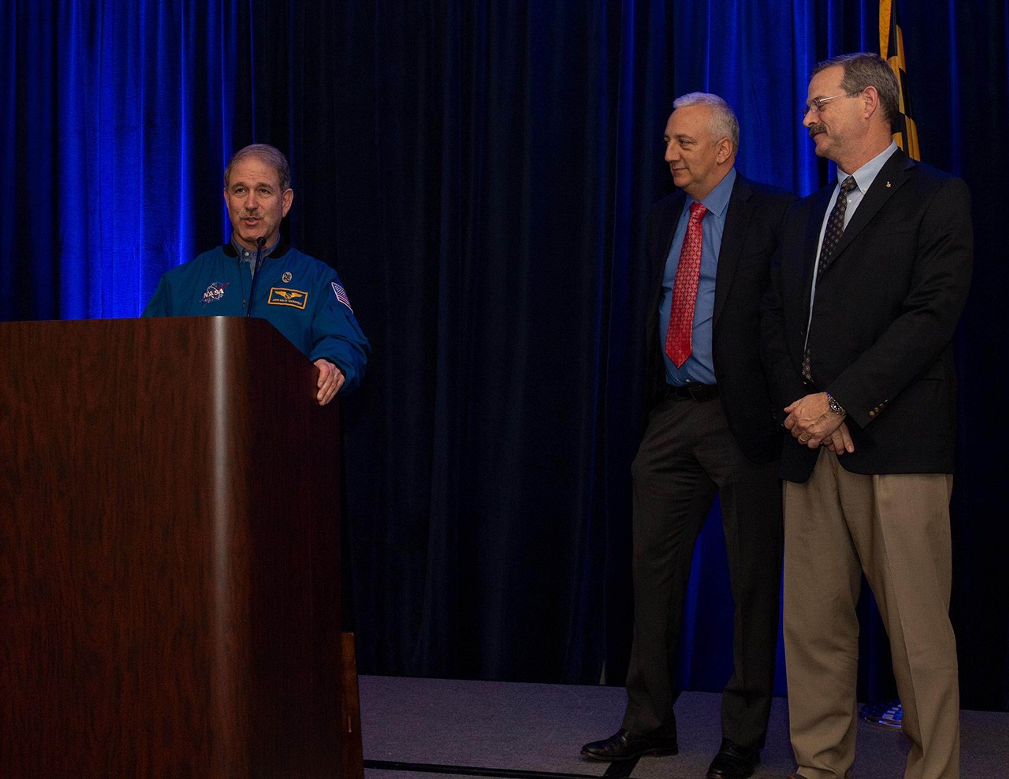 Grunsfeld (left) speaks at the podium while Massimino and Altman watch from the right.