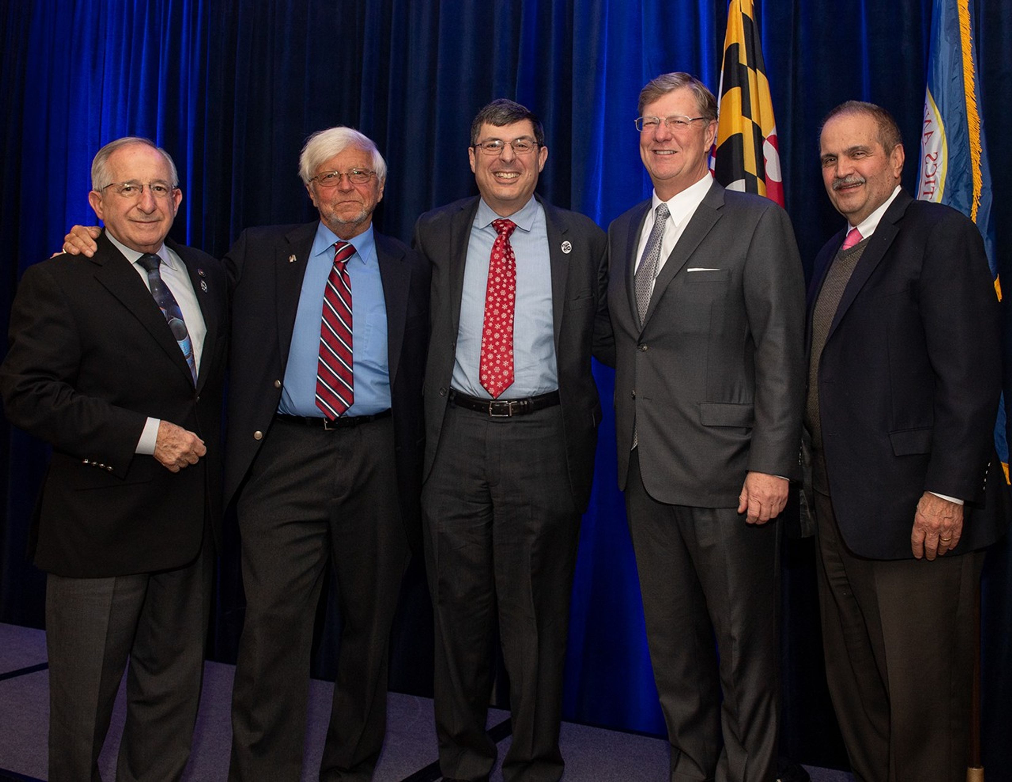 Five men stand in a row onstage, smiling at the camera.
