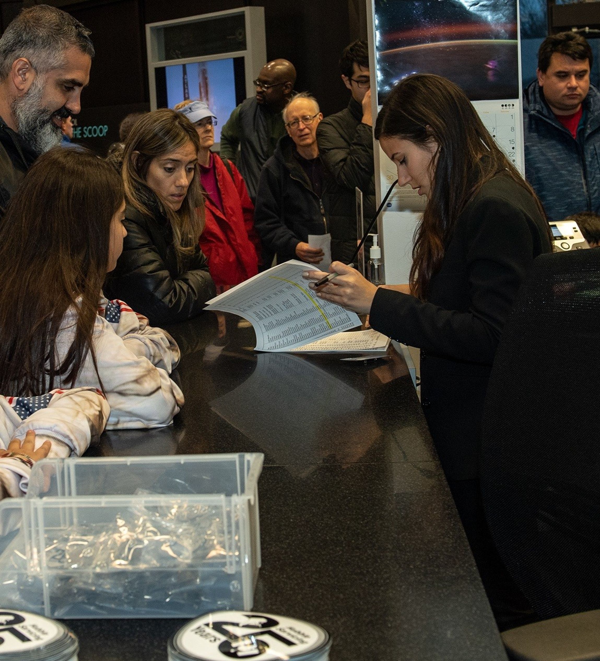 Erin Kisliuk (right) looks through a guest check-in list, while a family of visitors in the foreground on the left (a man, a woman, and two children) wait to be checked in. More visitors are visible in the background.
