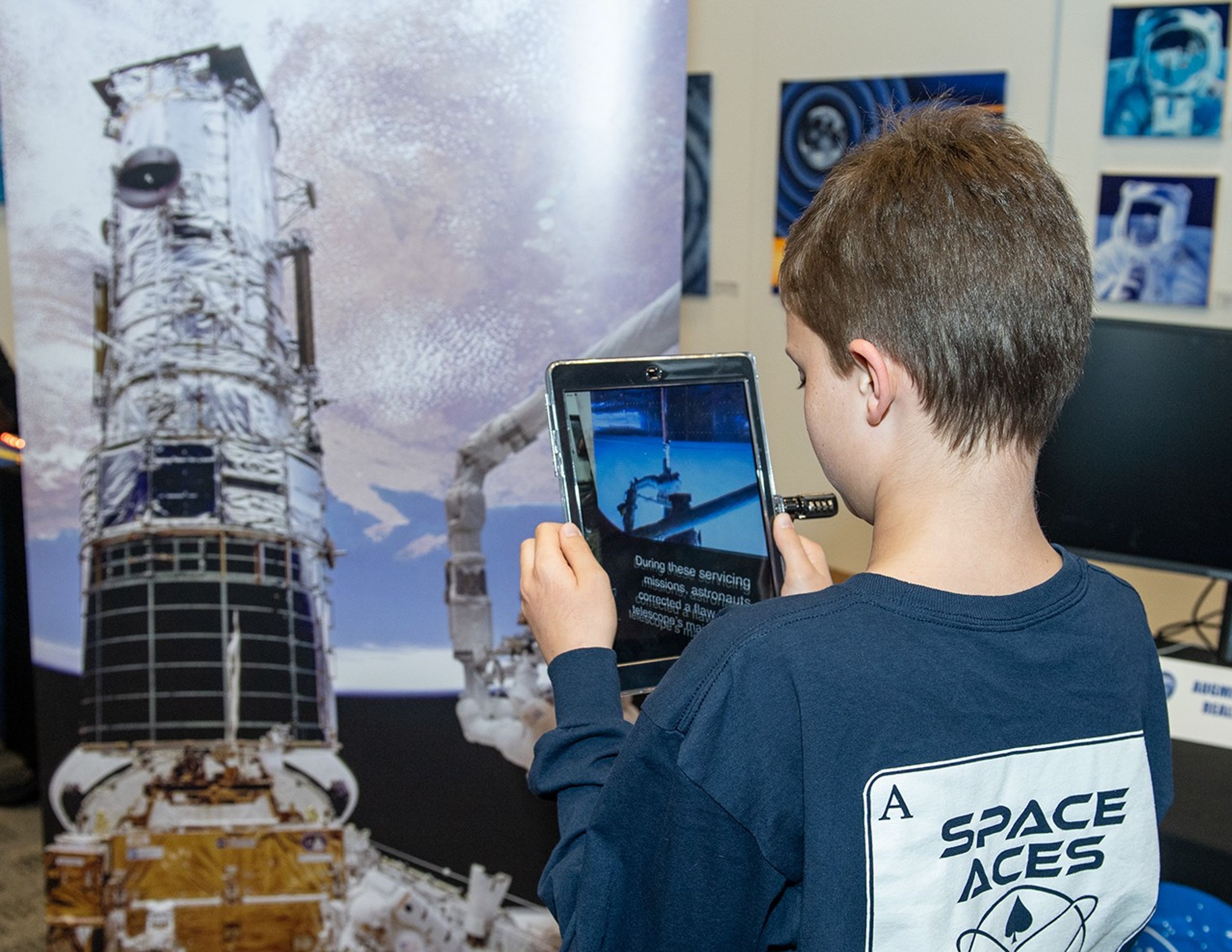 A boy, facing away from the camera on the right-hand side of the image, points an iPad at a Hubble servicing mission banner on the left side.