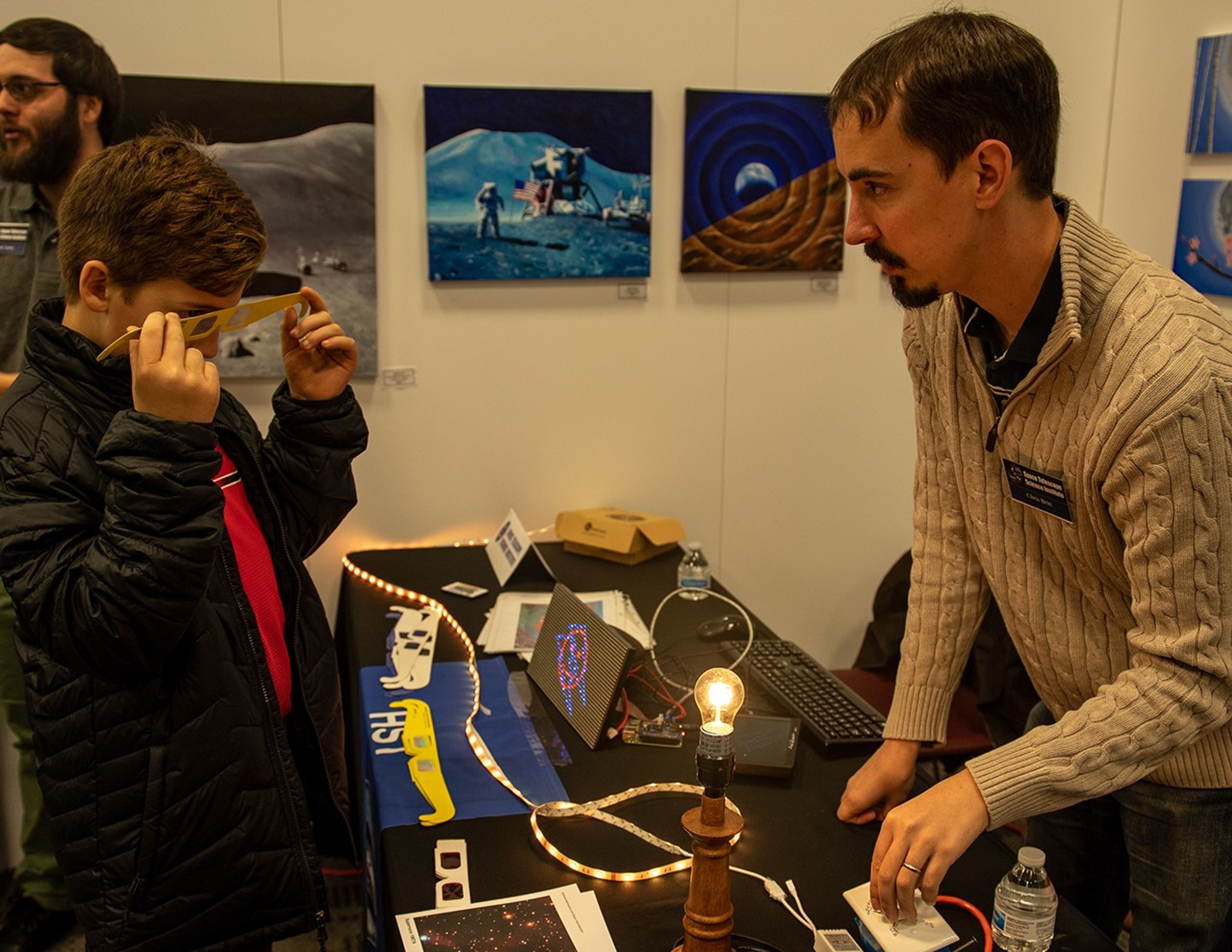 Chris Britt (right), standing behind the outreach table with interactive activities, speaks with a visitor, a boy who's holding a pair of yellow paper glasses with clear lenses. Another visitor is visible in the background on the left