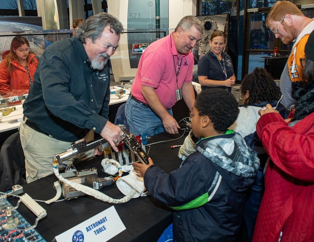 Two men (left and center) and one woman (right) stand behind the table with a black NASA tablecloth, showing astronaut tools to three visiting kids and their adult chaperone.