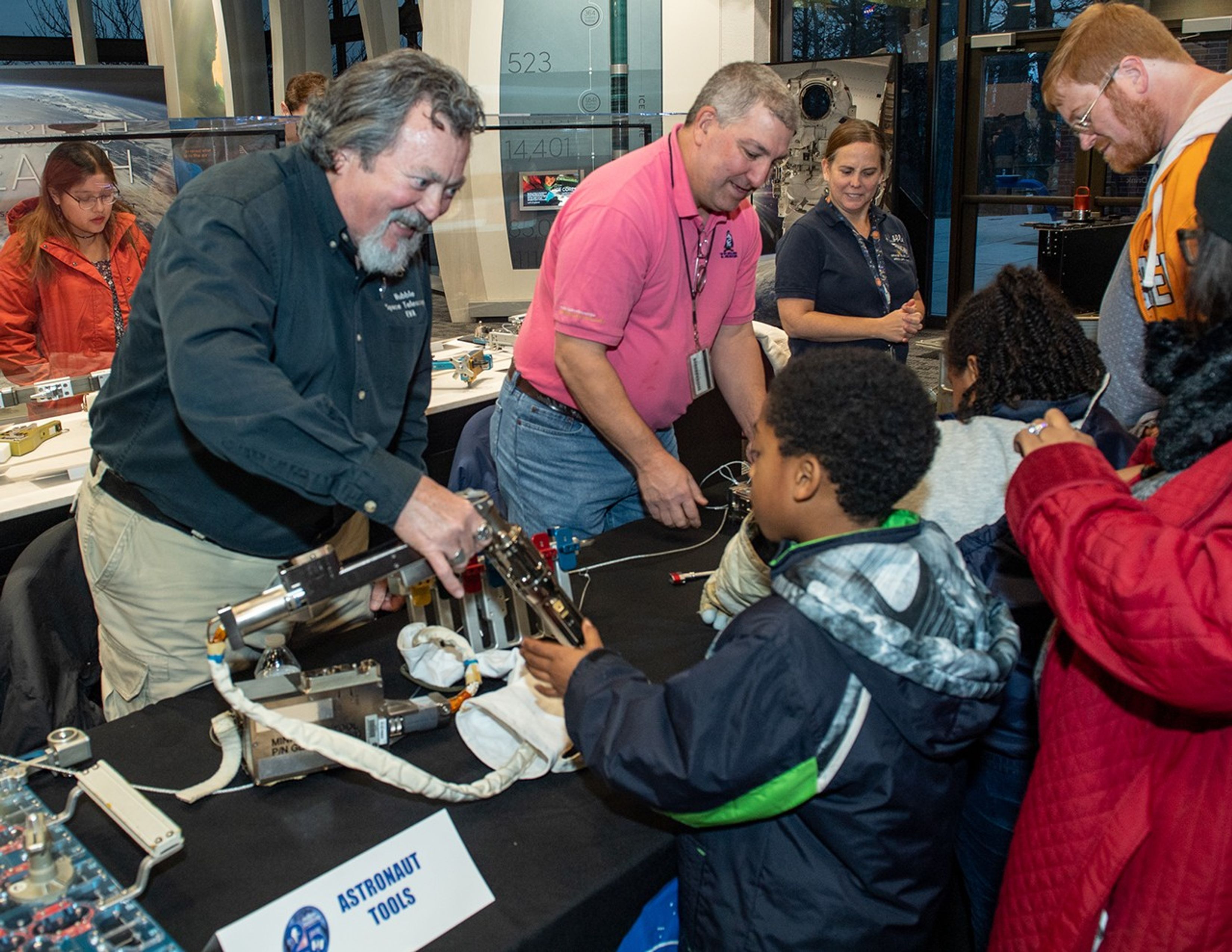 Two men (left and center) and one woman (right) stand behind the table with a black NASA tablecloth, showing astronaut tools to three visiting kids and their adult chaperone.