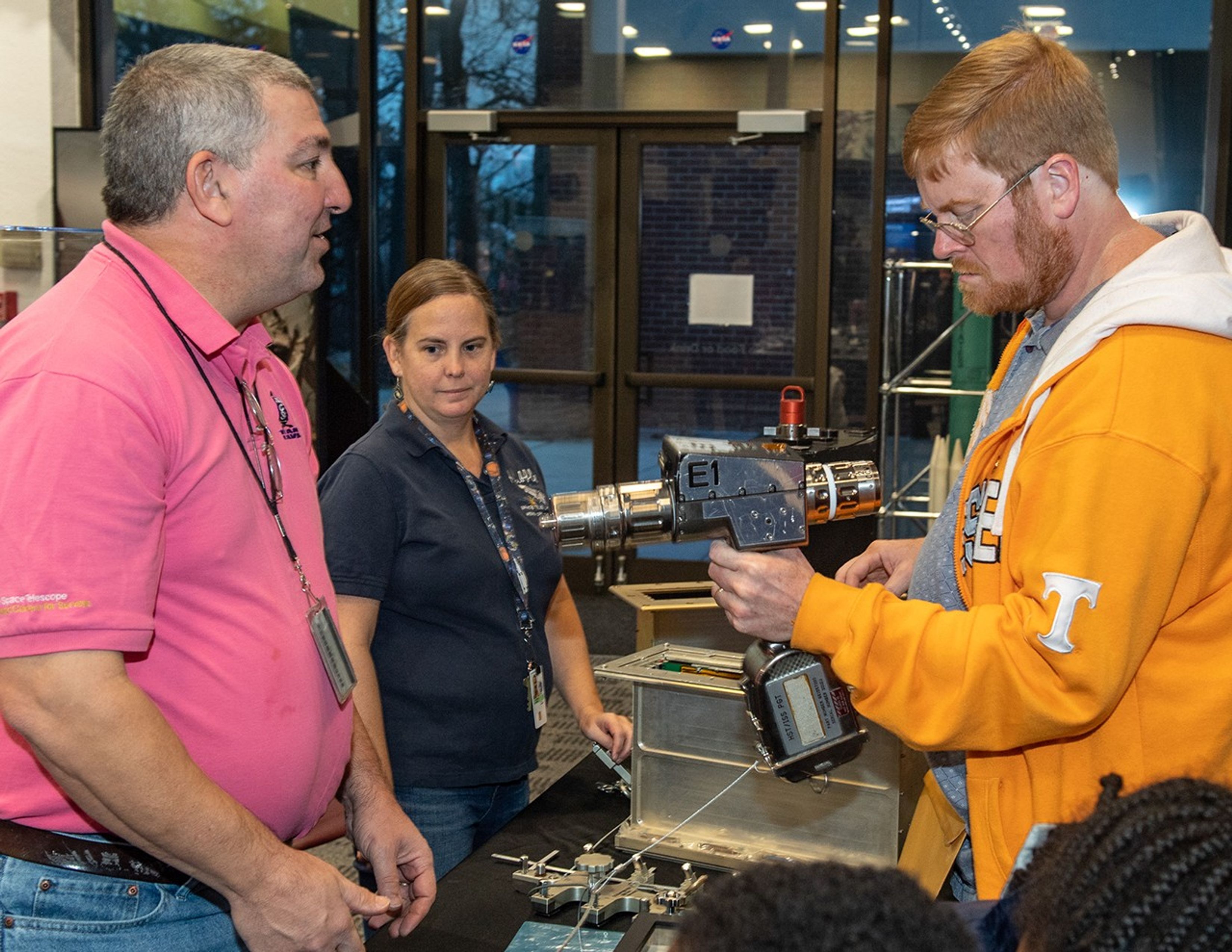 Steve (left) and Morgan (center), standing behind the table, allow a visitor (right) to hold an astronaut tool, with two children partially visible in the foreground.