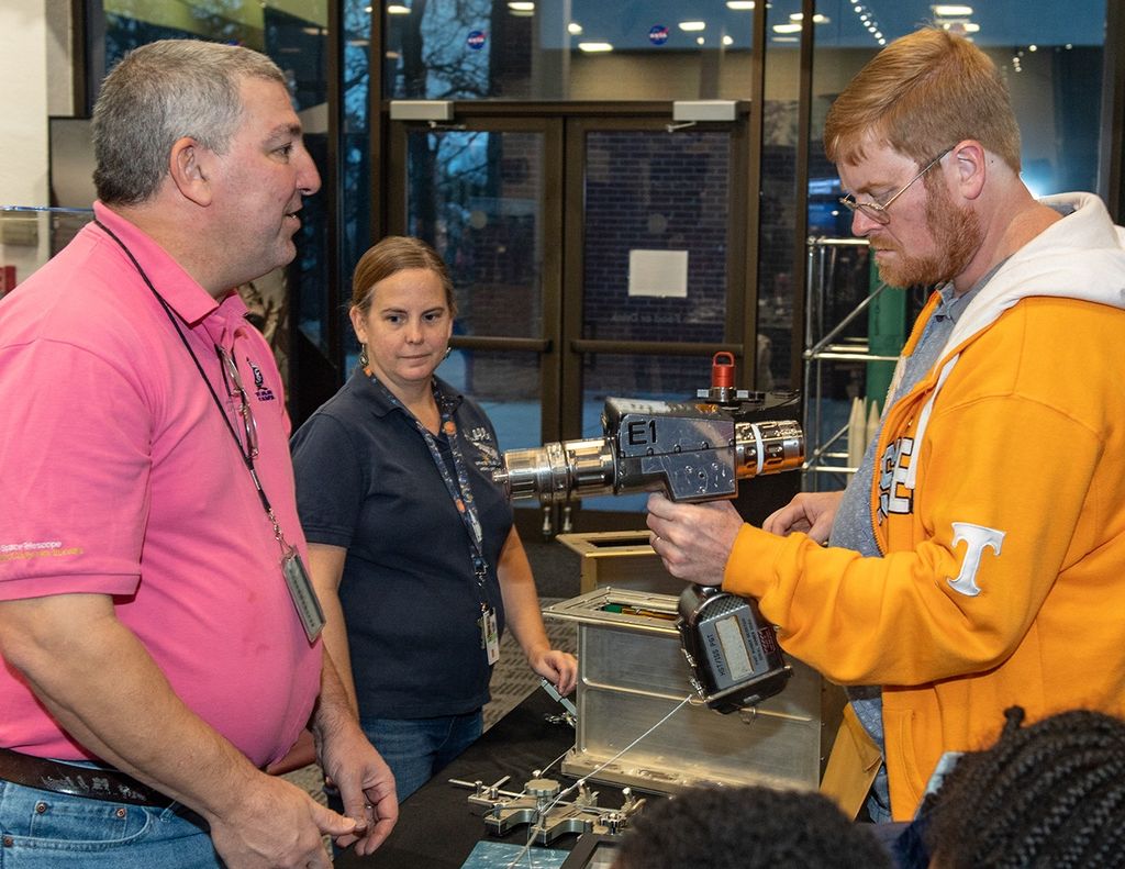 Steve (left) and Morgan (center), standing behind the table, allow a visitor (right) to hold an astronaut tool, with two children partially visible in the foreground.