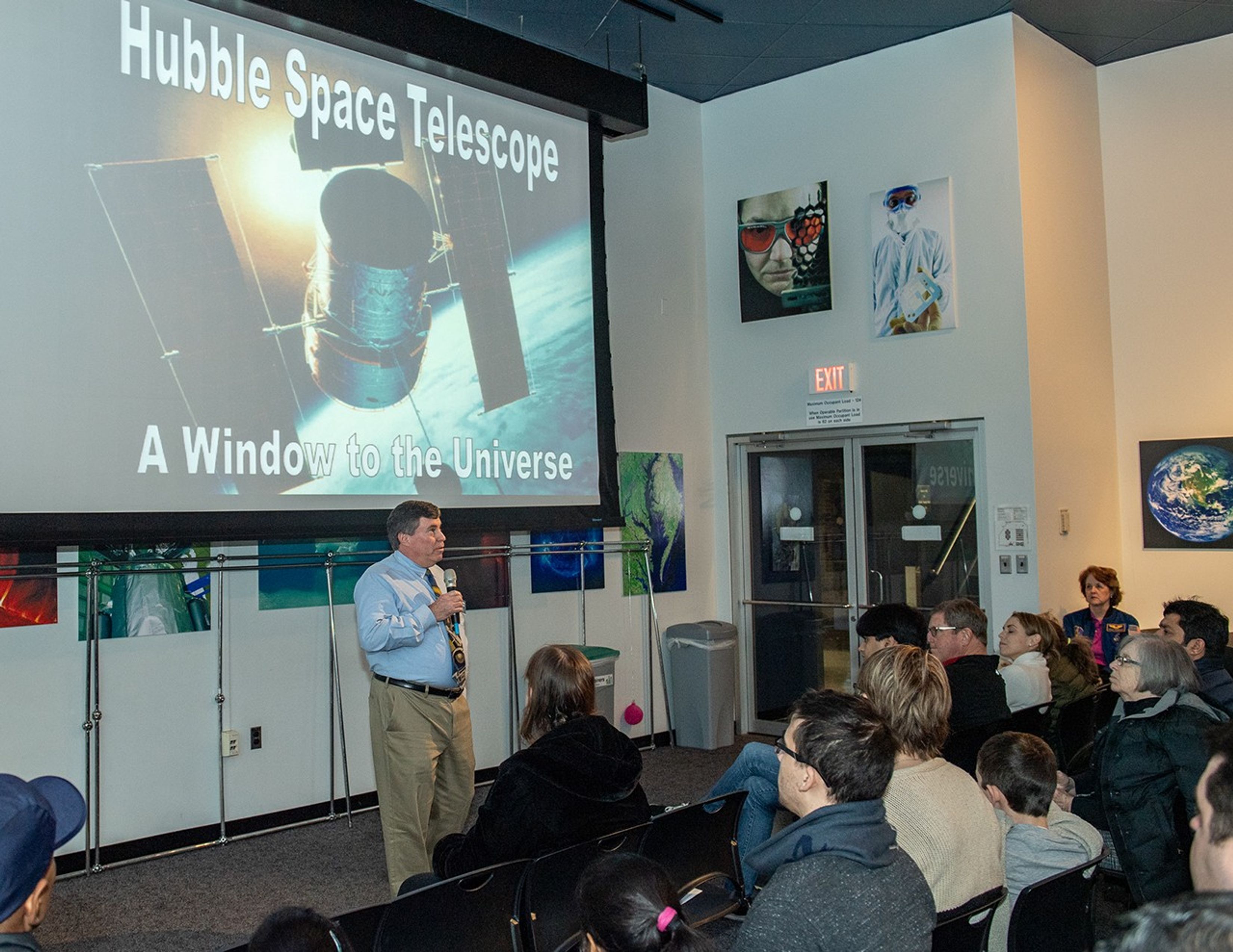 George Morrow (left) speaks with a microphone to a listening audience (right), with a picture of Hubble on the introductory slide on the screen above him.