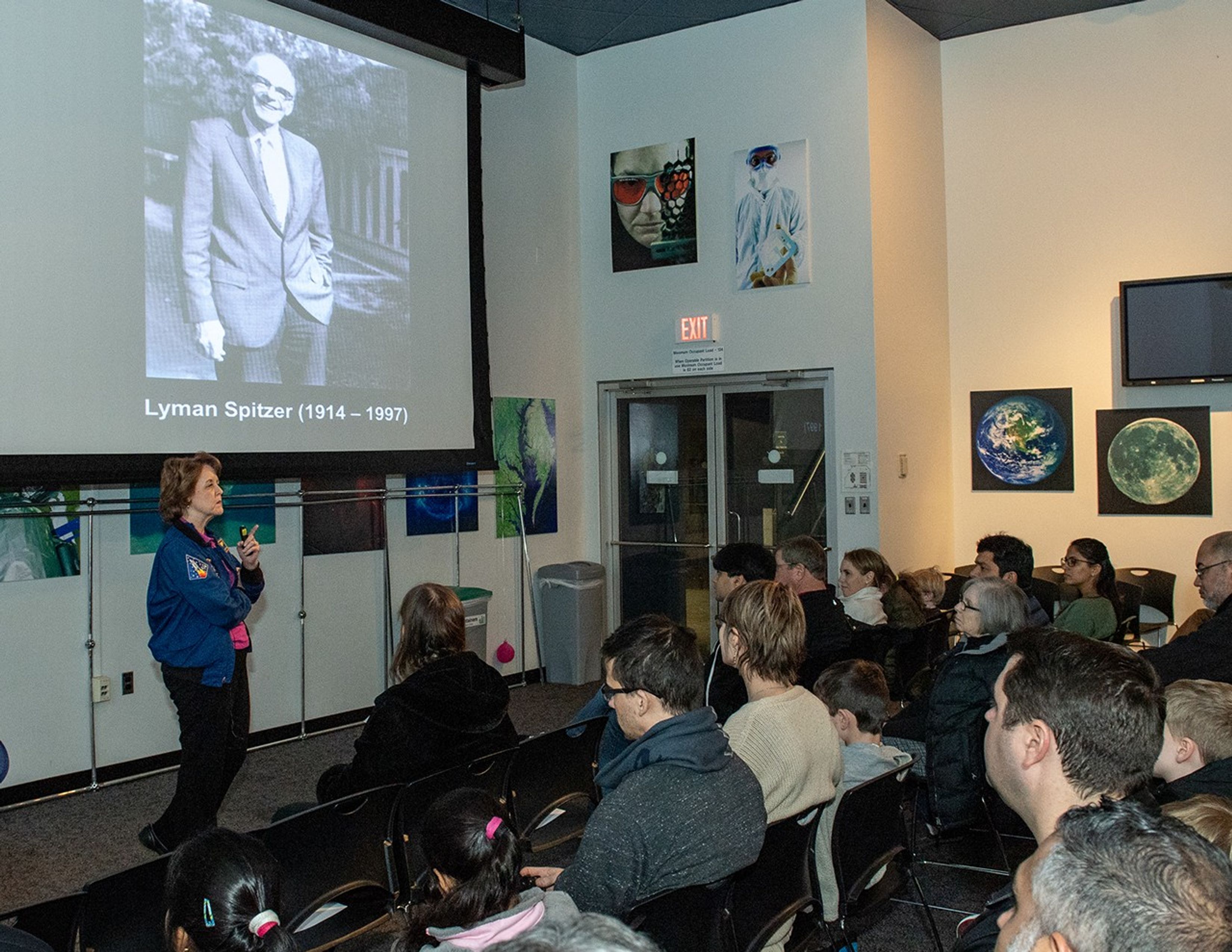 Kathy Thornton (left) speaks to a listening audience (right), with a picture of Lyman Spitzer slide on the screen above her.