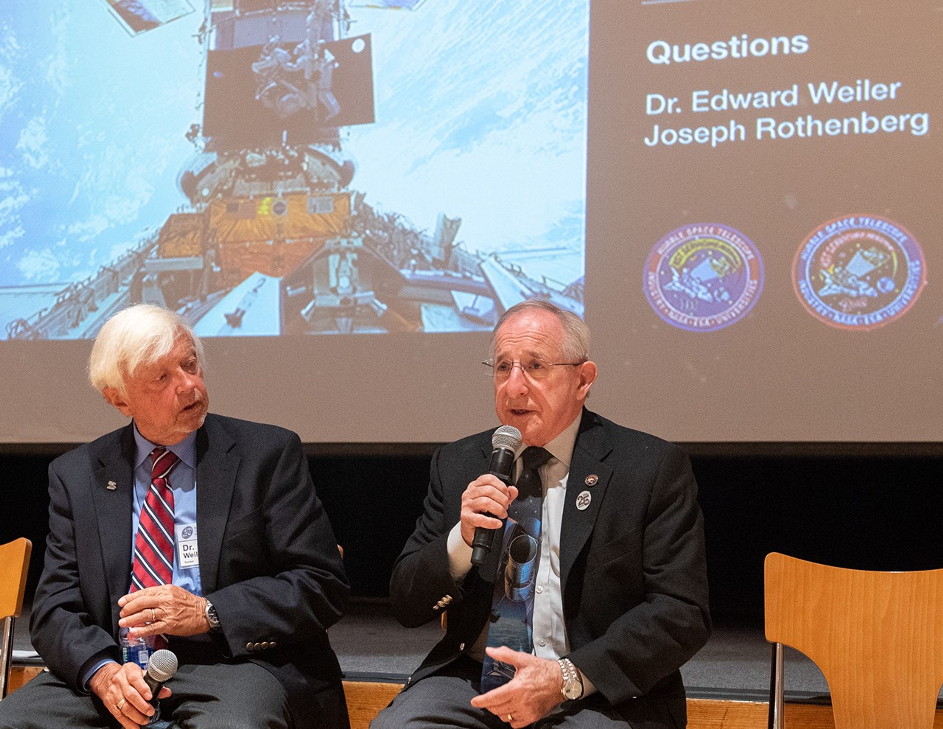 Joseph Rothenberg, right, speaks into a microphone to address the audience, with Ed Weiler, left, sitting next to him.