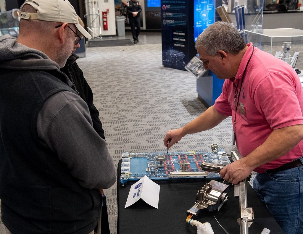 Arslanian (right), standing behind the table, demonstrates astronaut tools for two visitors (left).