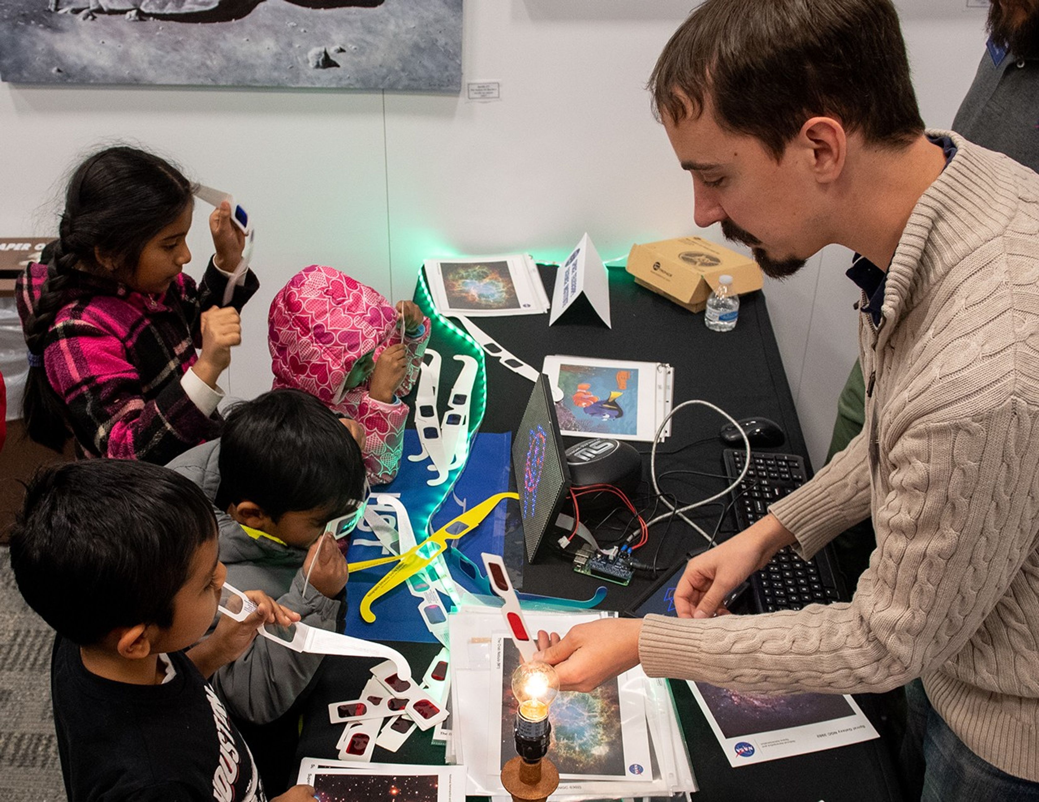 Britt (right), from behind the table, tells four kids (left) how to use the paper glasses with multi-colored lenses to look at the Hubble science image on the small screen.
