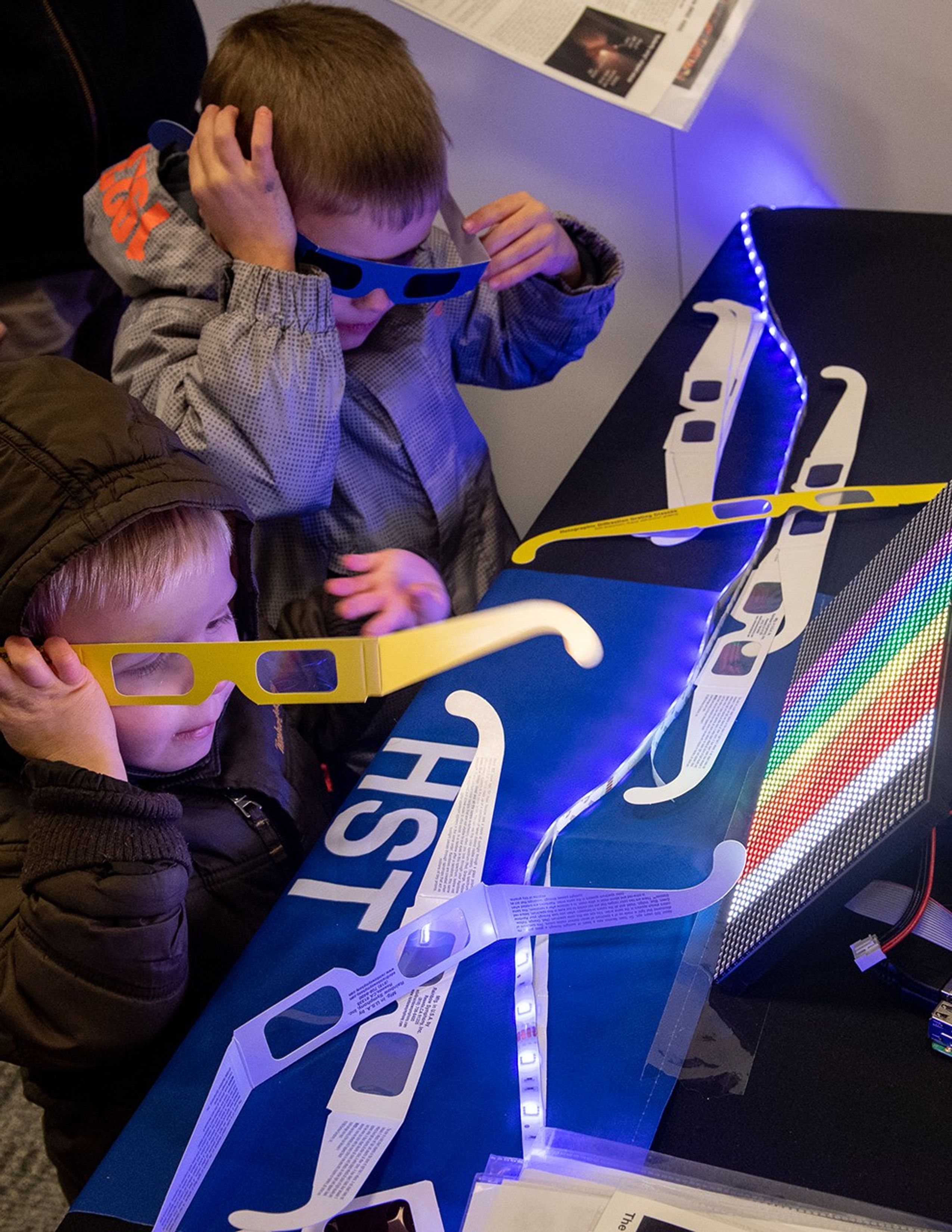 Two boys use paper glasses with clear diffraction lenses to look at a screen with rainbow colors on it.