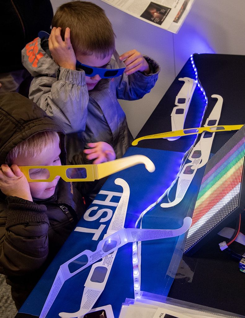 Two boys use paper glasses with clear diffraction lenses to look at a screen with rainbow colors on it.