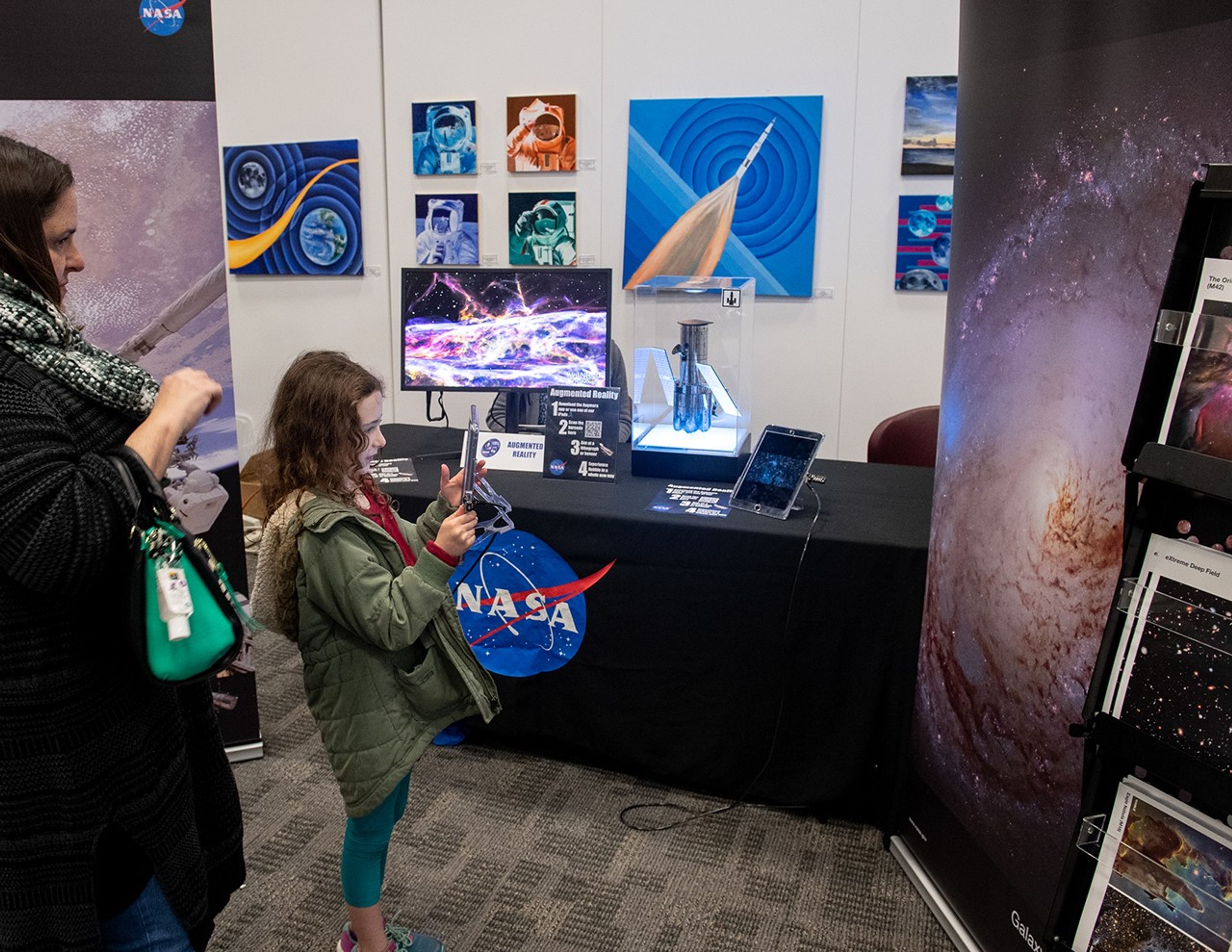 A girl (right) stands on the left side of the picture while pointing an iPad at a banner with a picture of a galaxy on the right side of the image. A table in the background has a black NASA tablecloth, a monitor on it with a Hubble image, and a scale model of Hubble. A chaperone (left) watches nearby.
