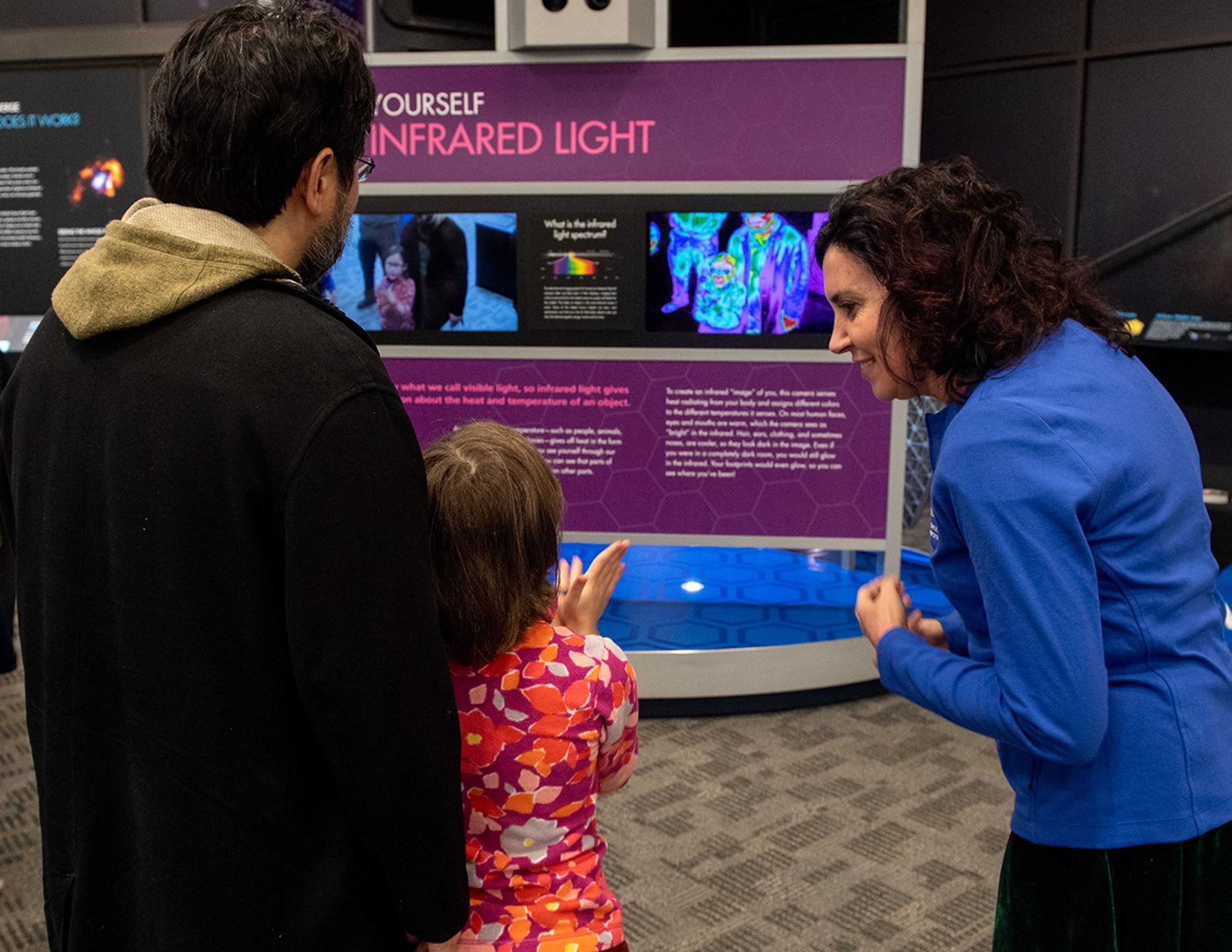 Begoña Vila, right, smiles while speaking to a child, center, who stands with a chaperone, left, while using the Webb Infrared Camera exhibit.