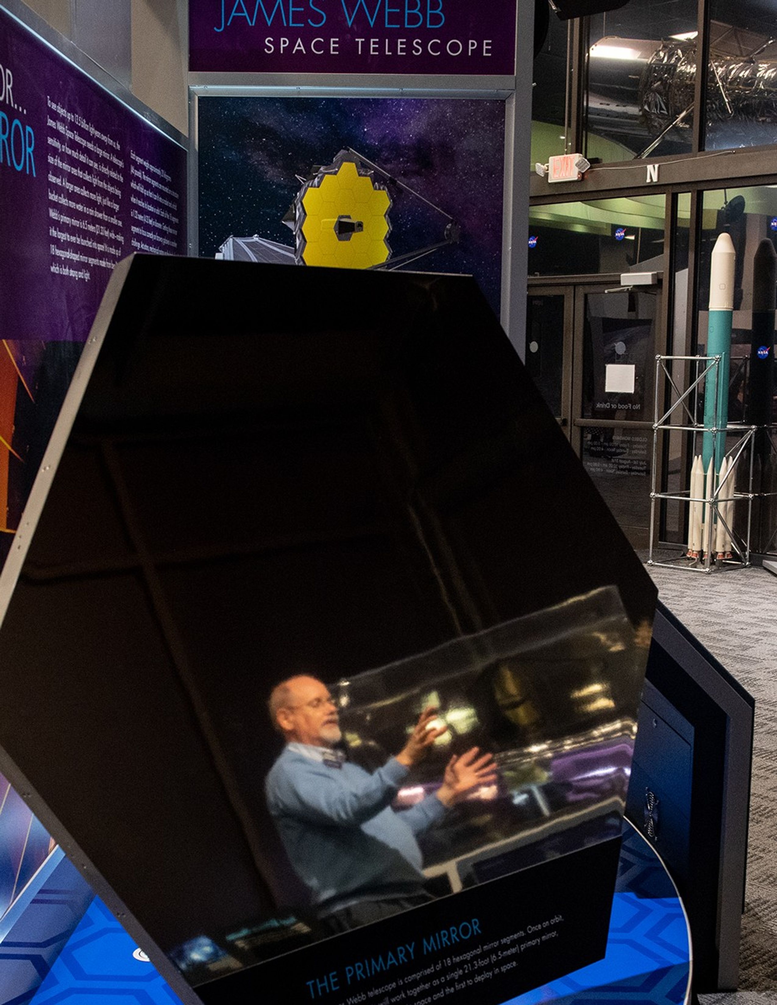 Randy Kimble and the model of the Webb Space Telescope that he's explaining are both reflected in the hexagonal Webb sample mirror segment.